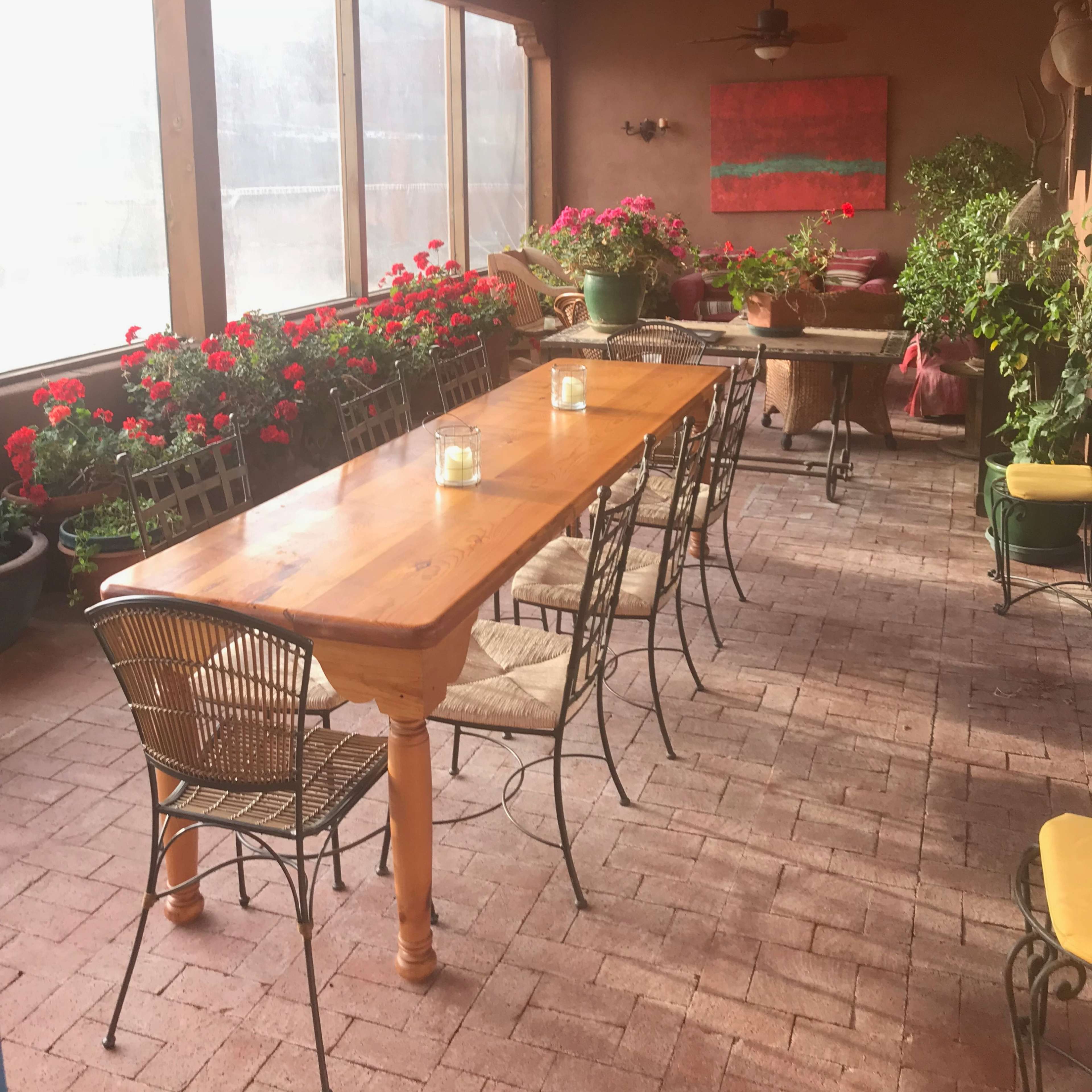 A spacious patio featuring a long wooden dining table surrounded by chairs, with flower-filled planters and a painting on the wall.