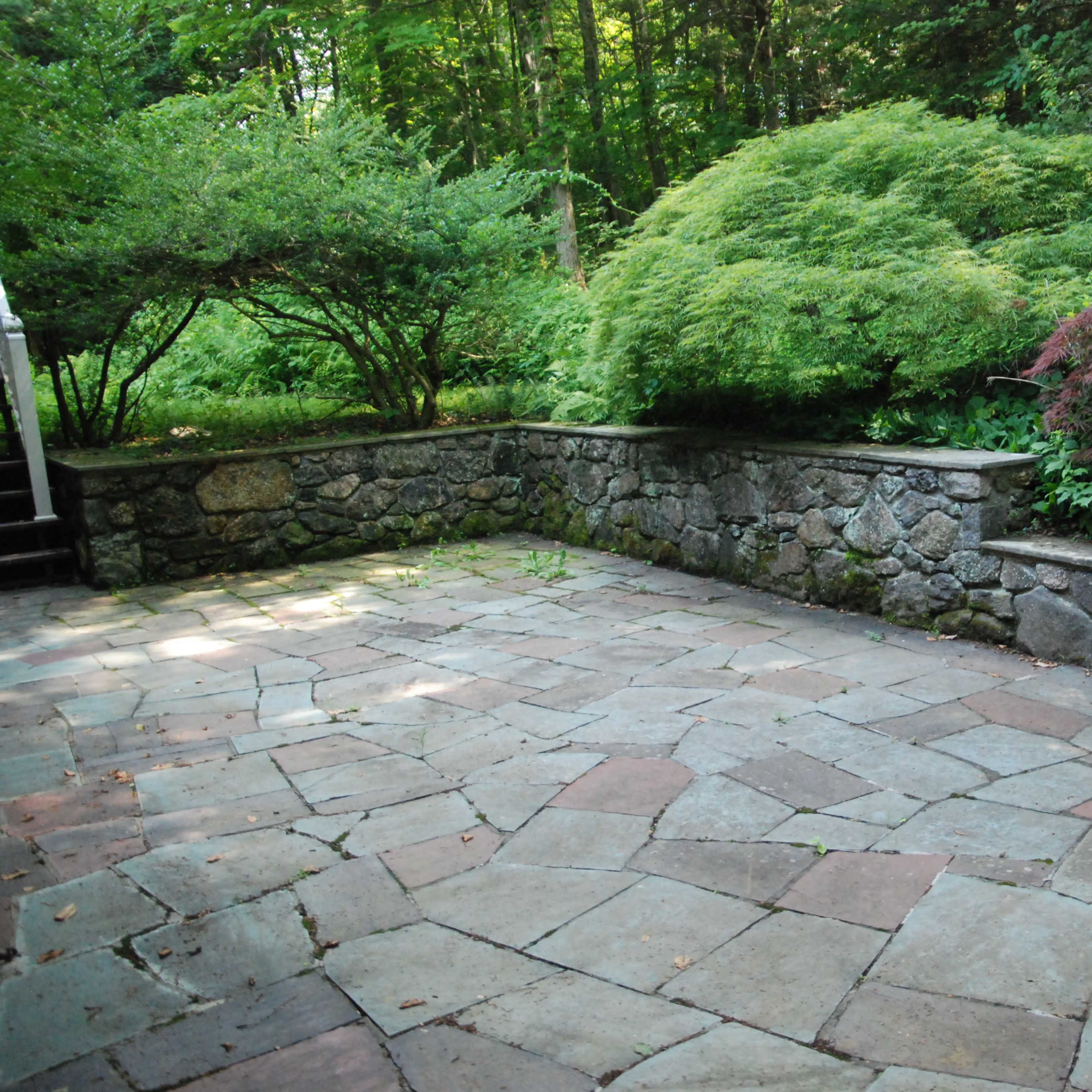 A stone patio is surrounded by lush greenery and shrubbery along a low stone wall.