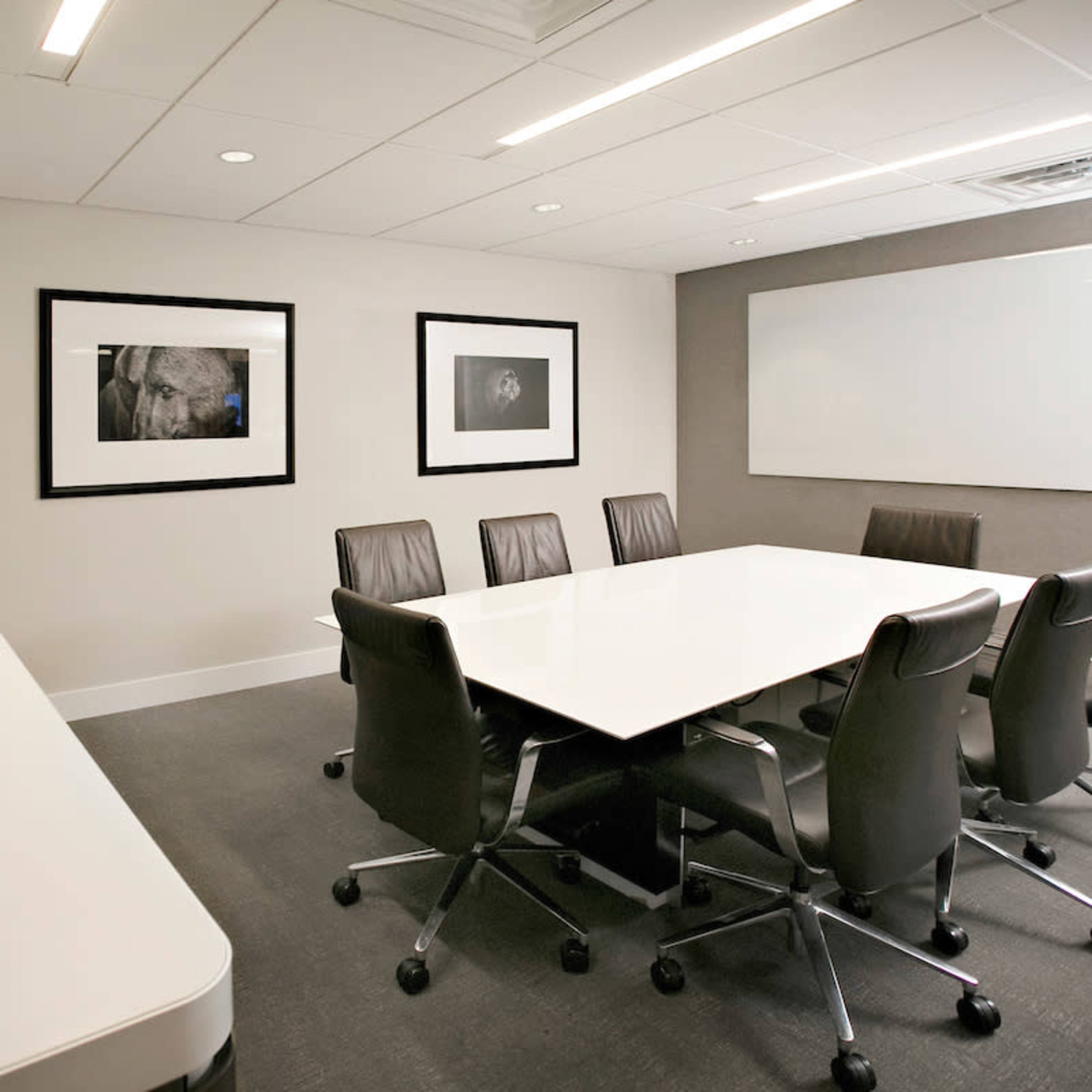 A modern conference room features a large white table surrounded by black leather chairs, with framed photographs on the walls and a flat-screen TV.