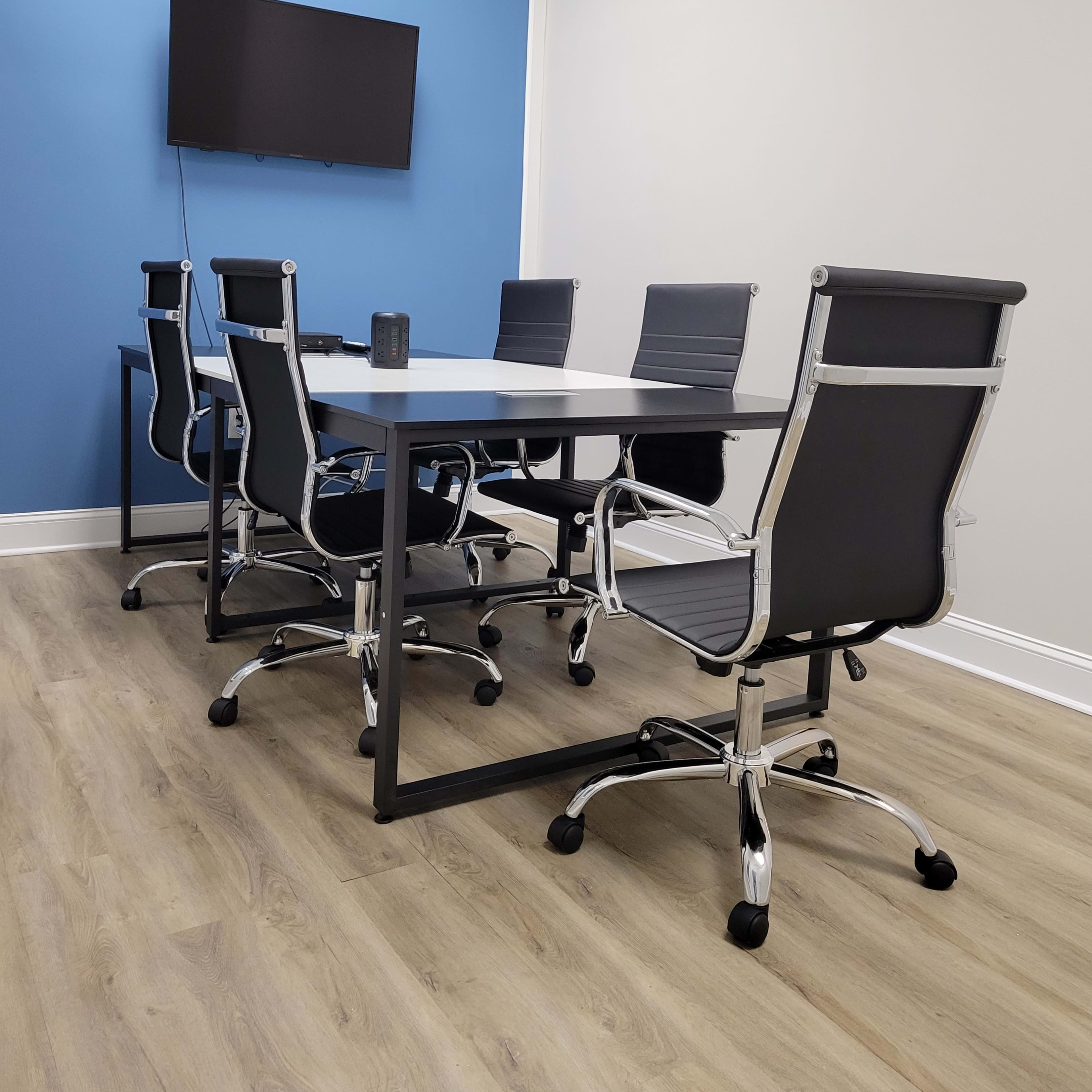 A modern conference room features a rectangular table surrounded by six rolling chairs and a wall-mounted television.