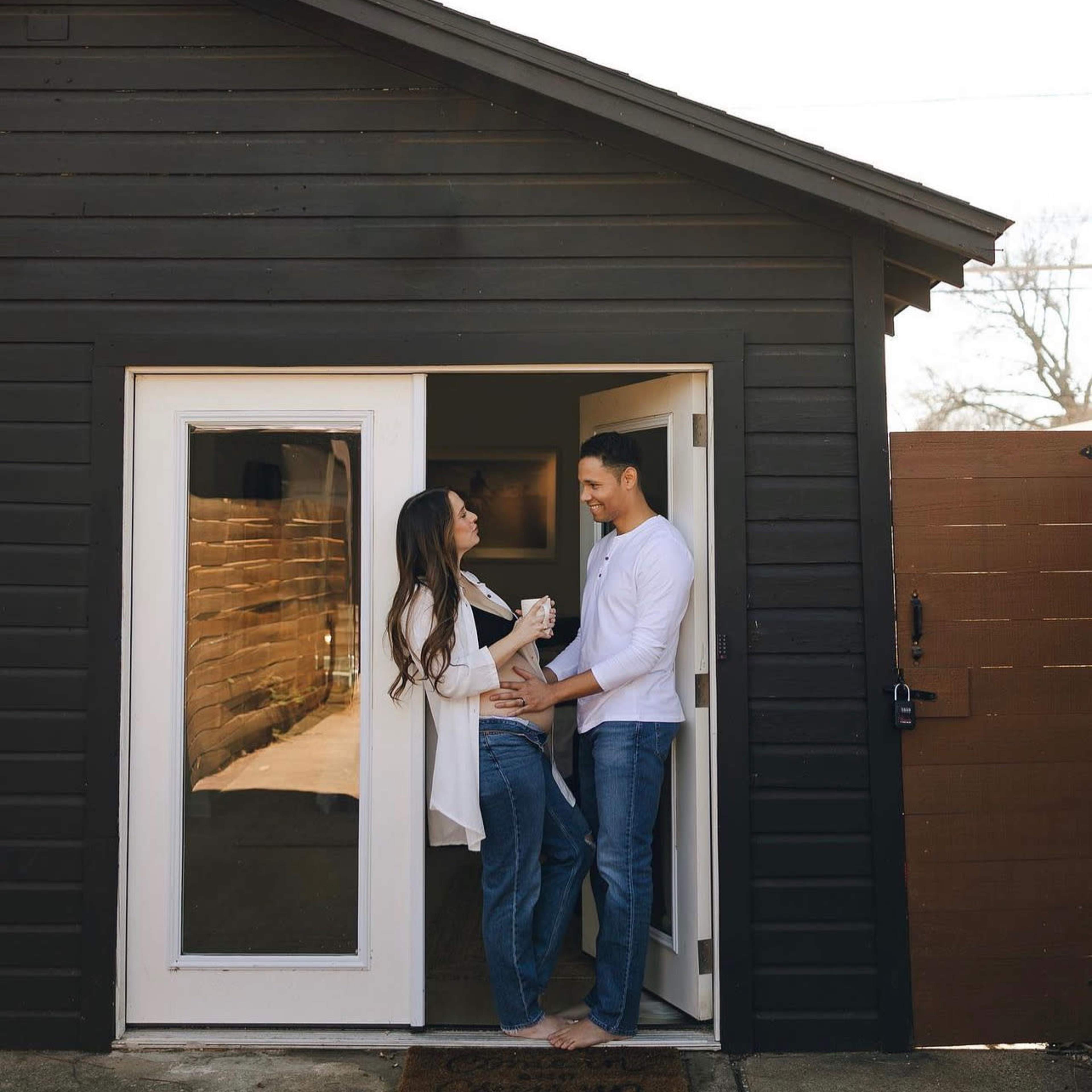 A couple stands playfully in a doorway of a black wooden house, enjoying each other's company.