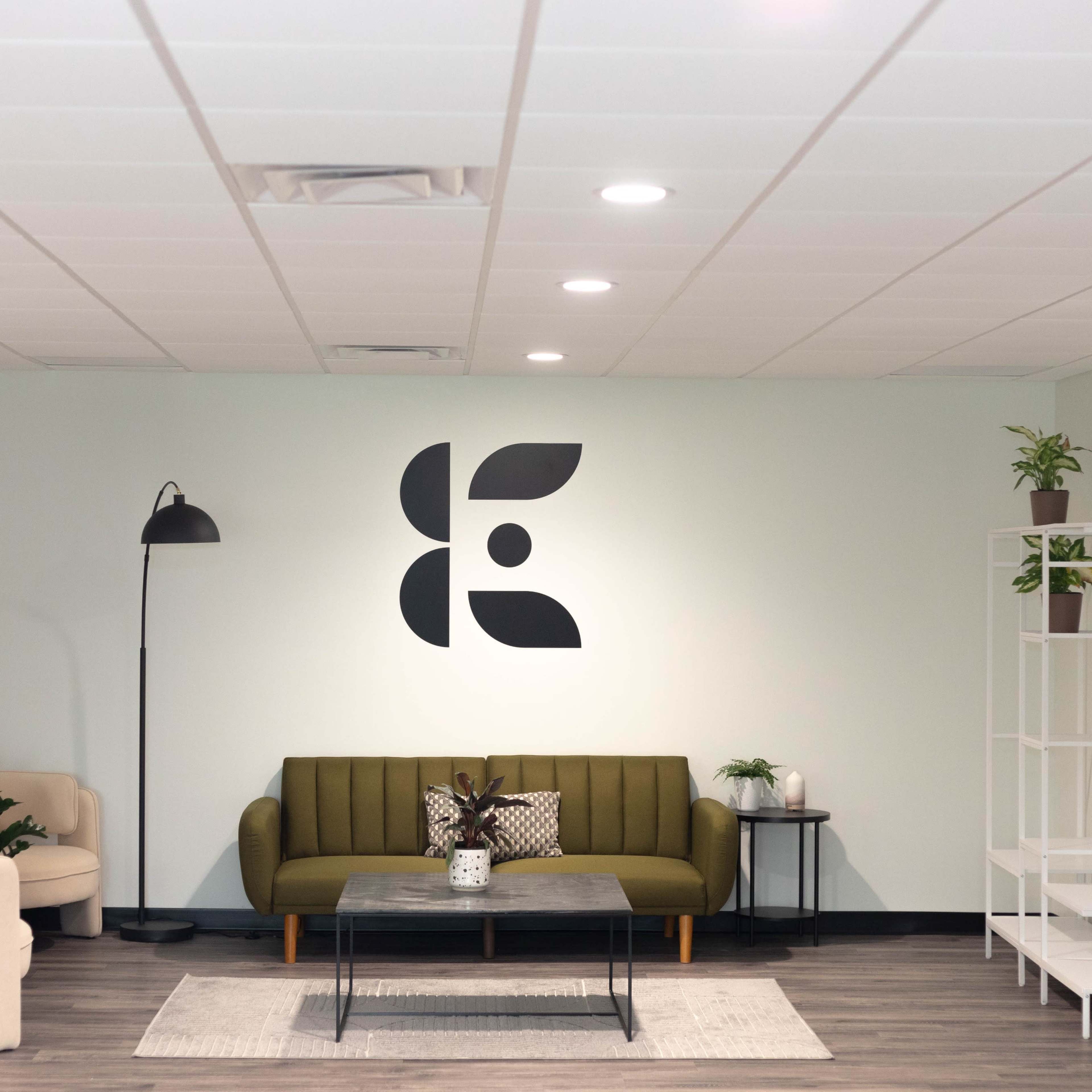A modern waiting area features a green couch, a white armchair, a coffee table, and a shelf with plants against a light blue wall displaying a large black logo.