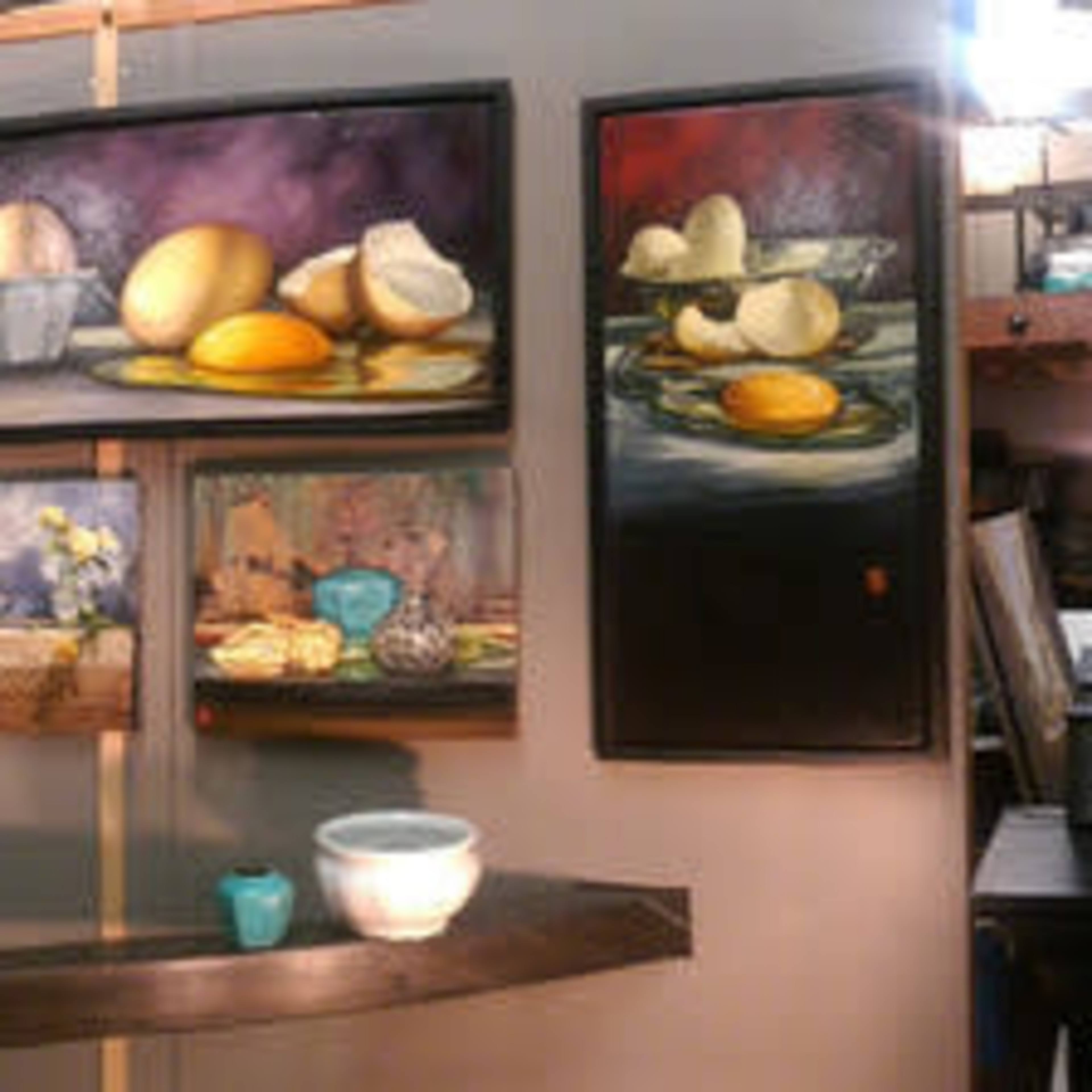 An art gallery featuring a variety of paintings displayed on walls, with artists' workstations and supplies visible in the background.