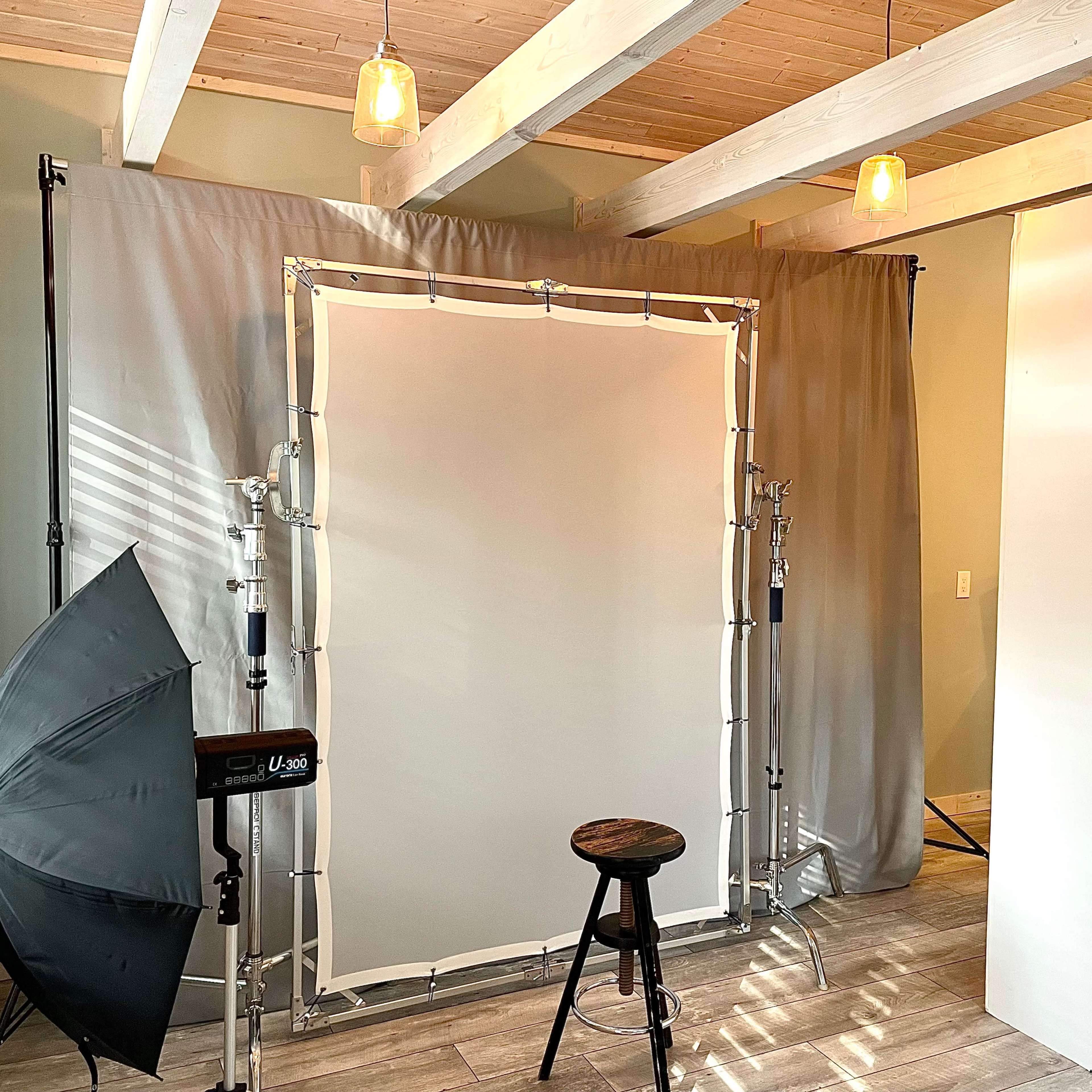 A photography setup featuring a backdrop with lighting equipment and a stool in a room with wooden beams and a light-colored floor.