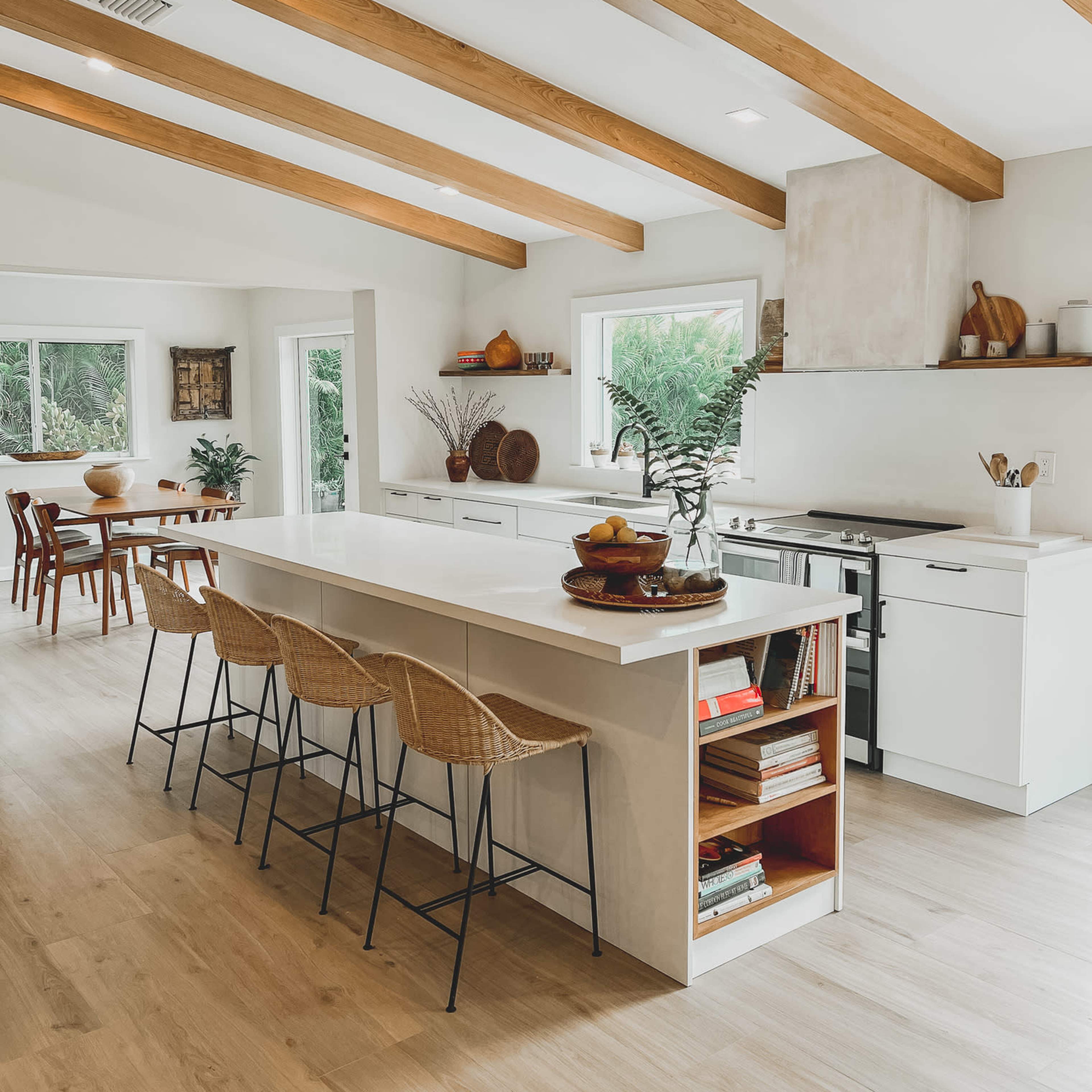 A modern kitchen features a long island with bar stools, white cabinetry, and exposed wooden beams overhead, along with a dining area in the background.