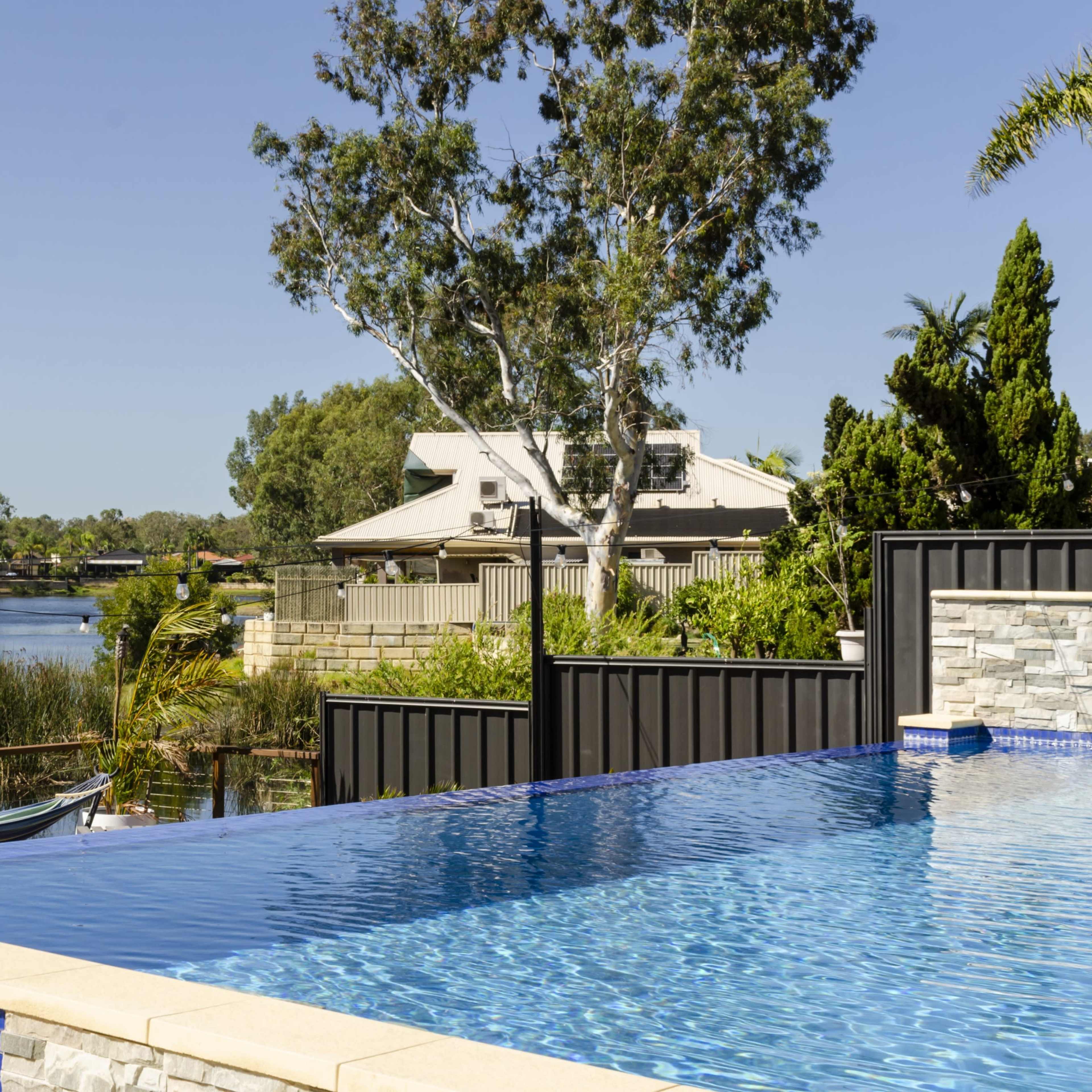 A modern infinity pool overlooks a calm river framed by lush greenery and residential buildings.