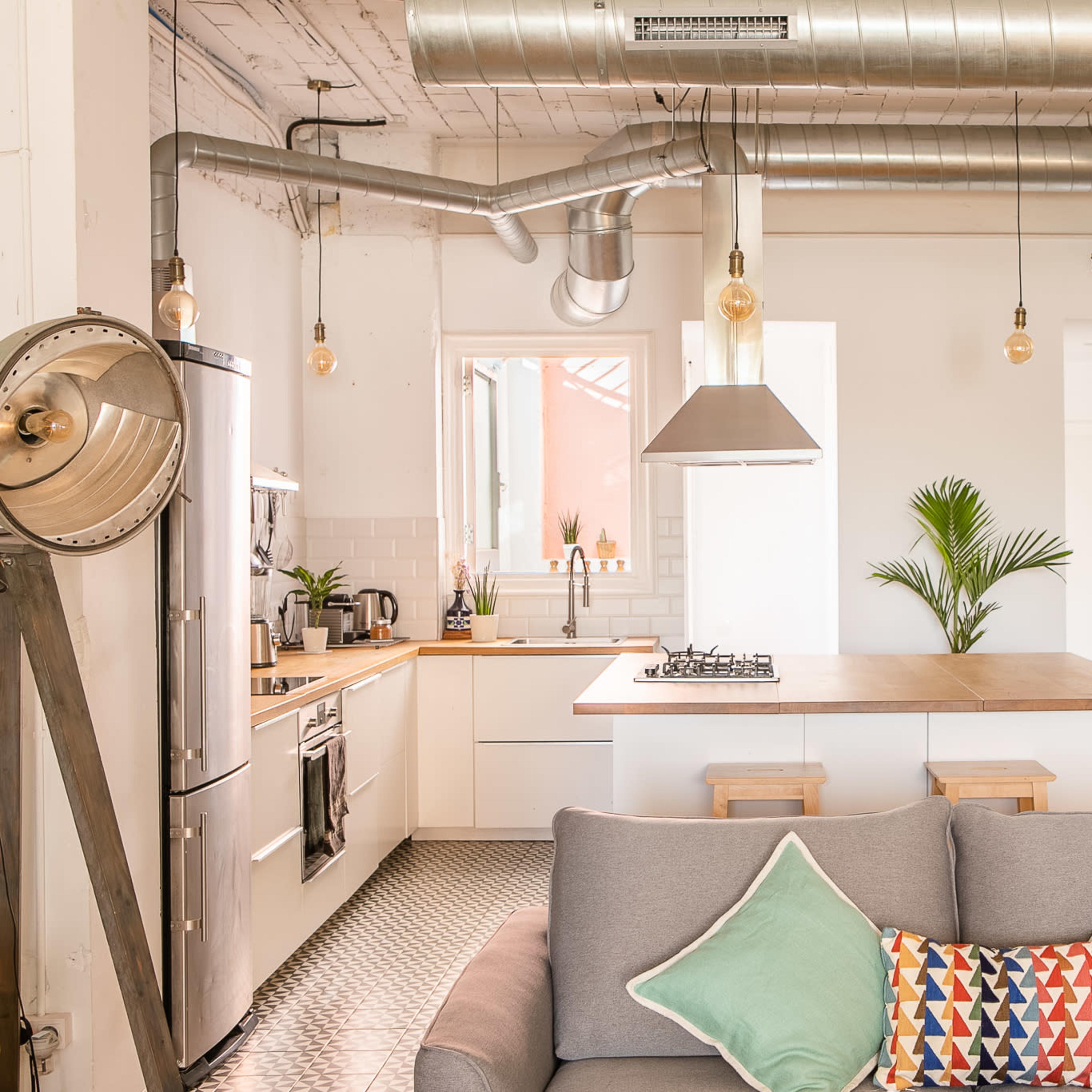 A modern kitchen and living area with exposed metal ductwork, a central island, and a sofa adorned with colorful cushions.