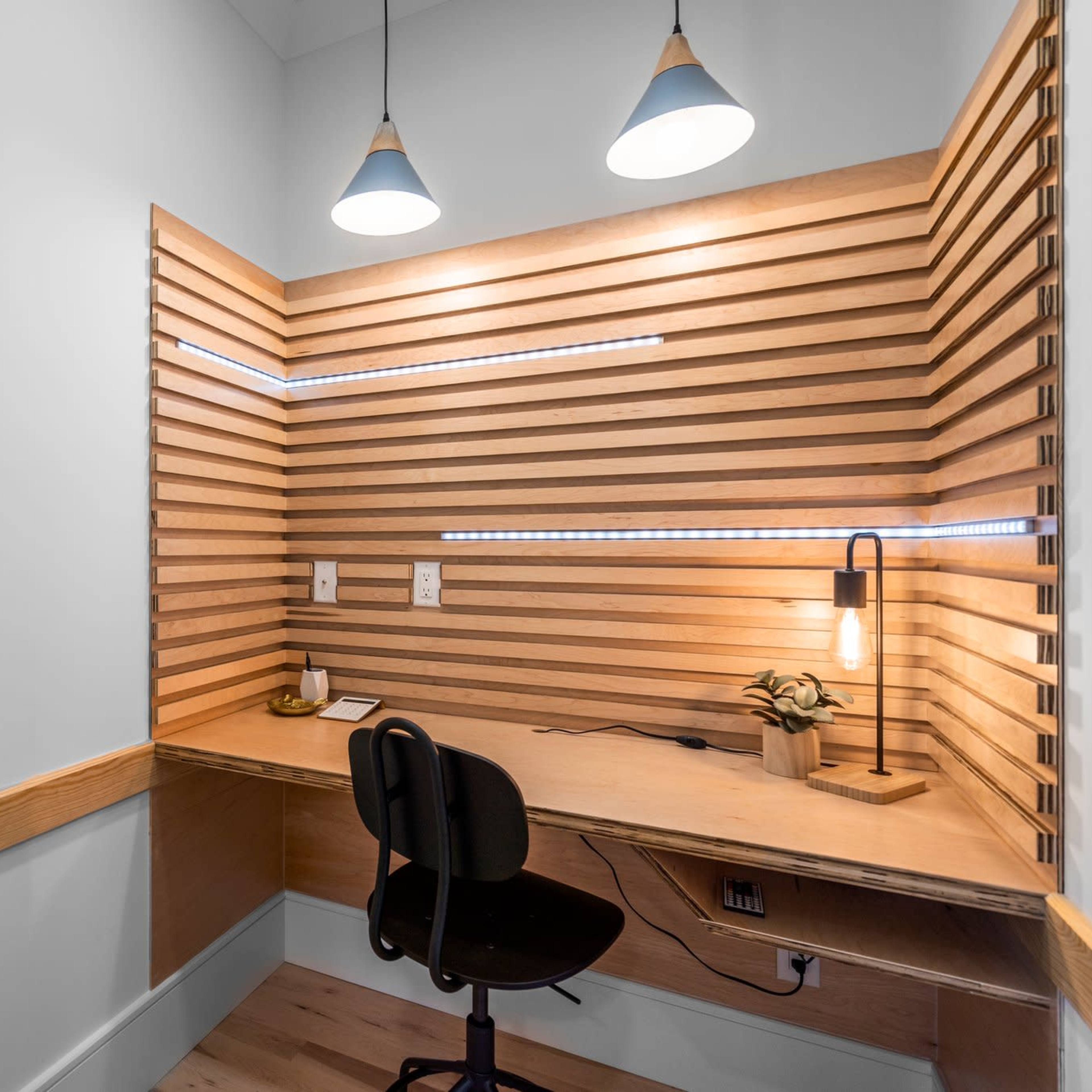 A minimalistic workspace features a wooden desk with a chair, surrounded by a slatted wooden wall and illuminated by pendant lights.