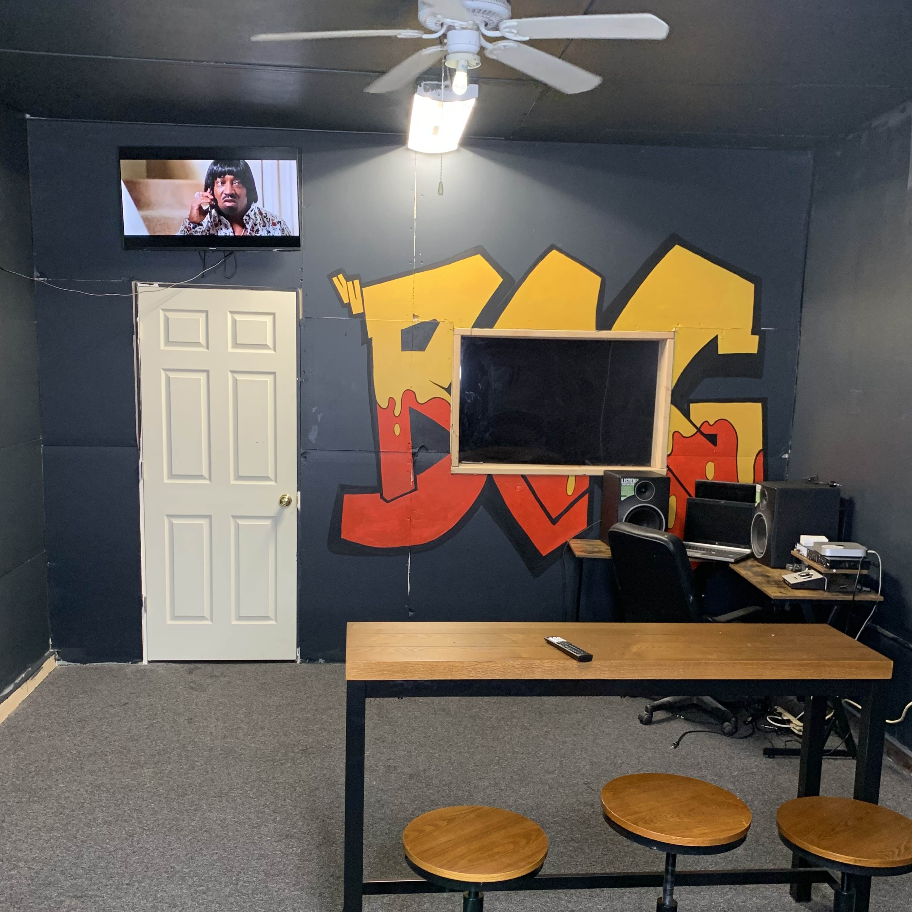 A room with a large graffiti-style "BIG" on the wall, a table with three stools, a computer setup, and a television mounted on the wall.