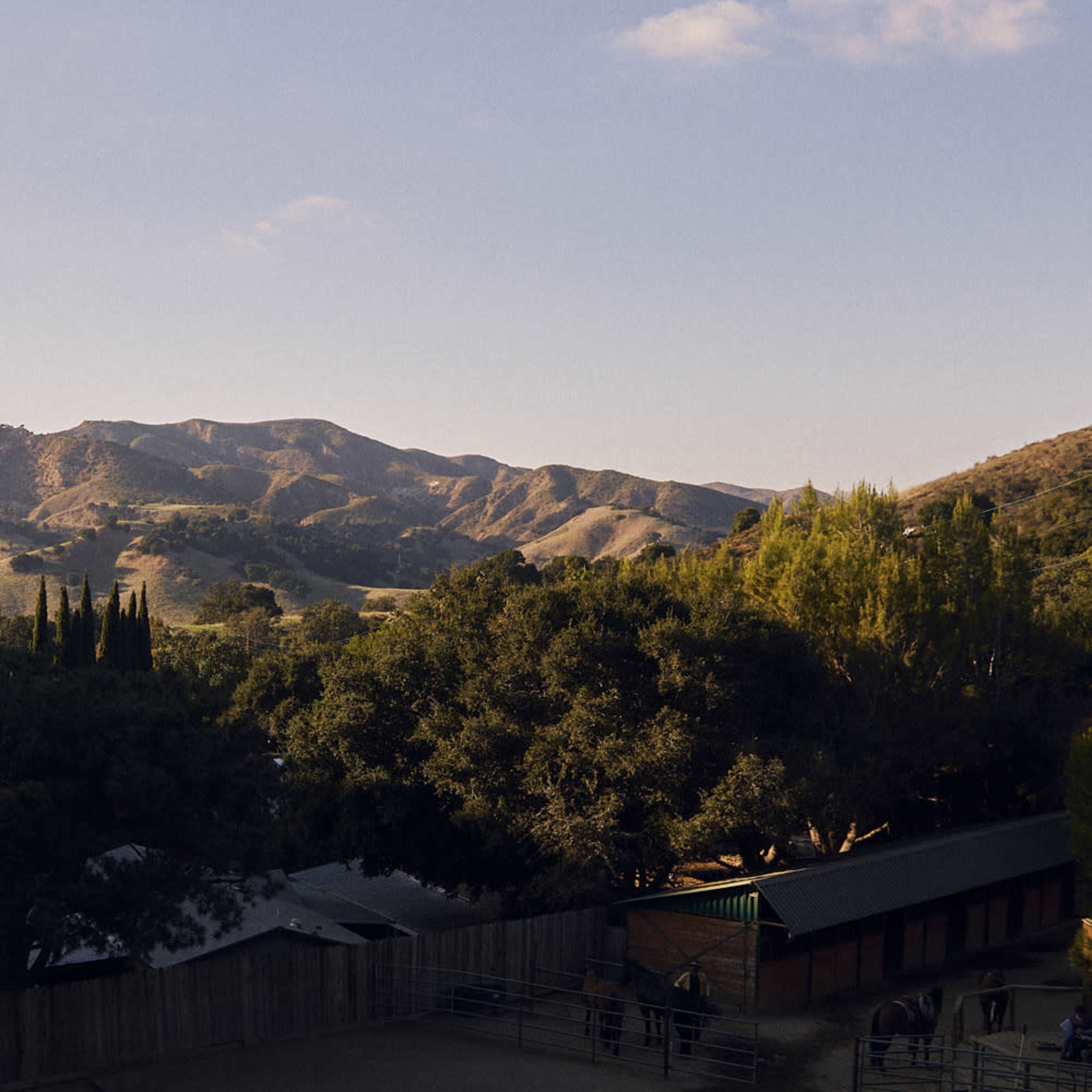 A rural landscape with rolling hills, dense trees, and a stable building in the foreground under a clear sky.