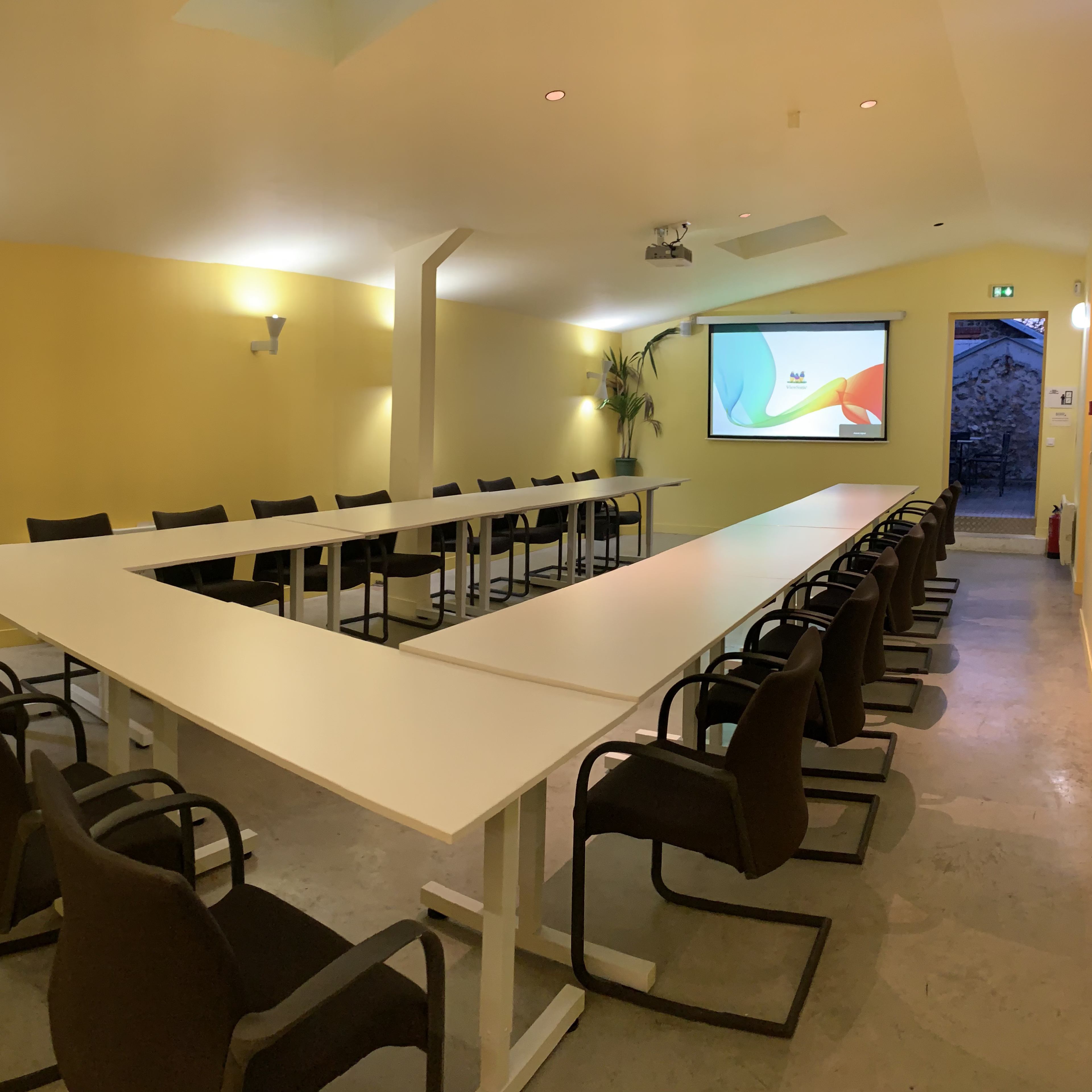 A modern conference room with a long, white table, black chairs, and a projector screen on a yellow wall.