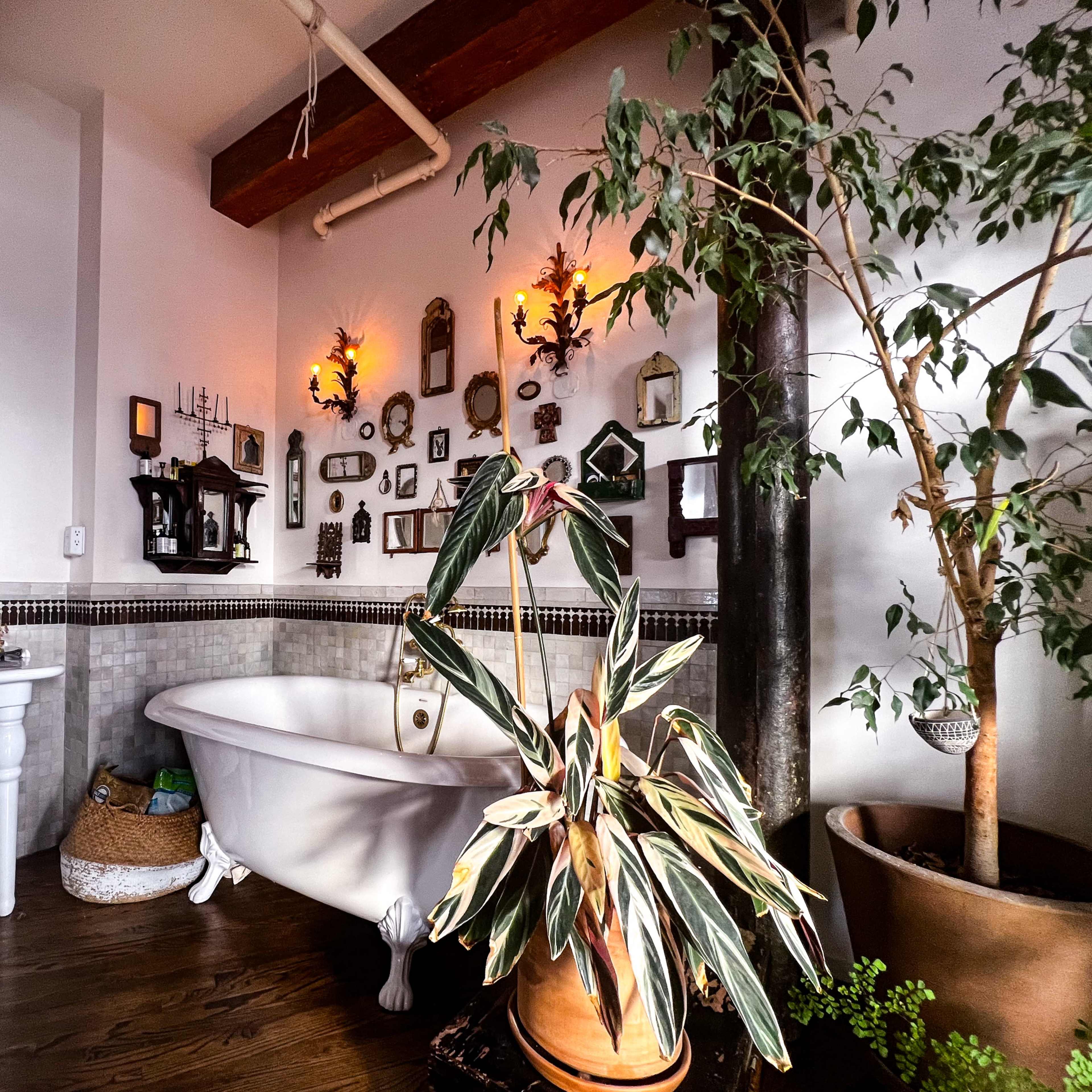 A bathroom with a freestanding bathtub, a large mirror, decorative wall-mounted items, and a potted plant beside a column.