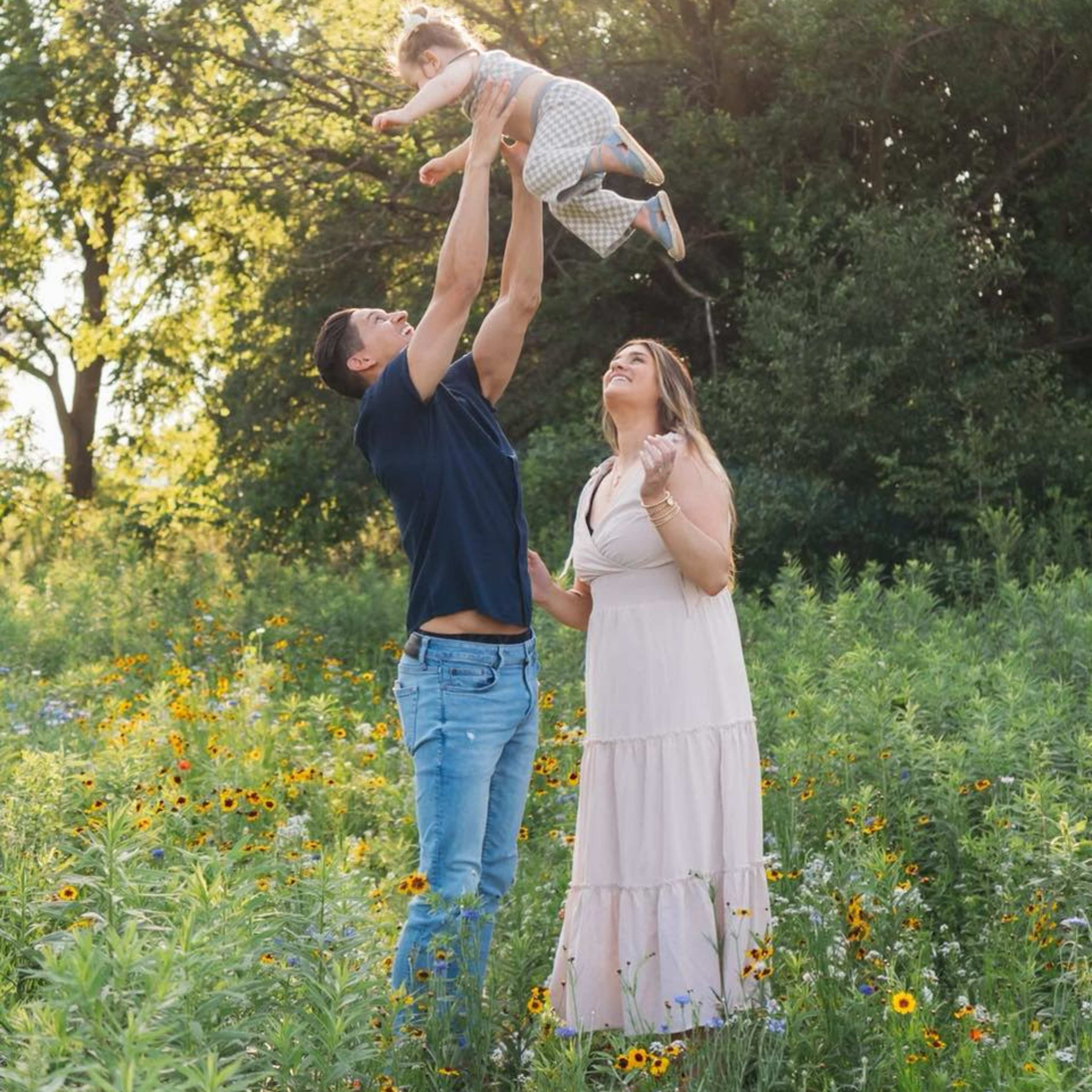 A man lifts a child above his head while a woman smiles in a field of wildflowers under a bright sun.