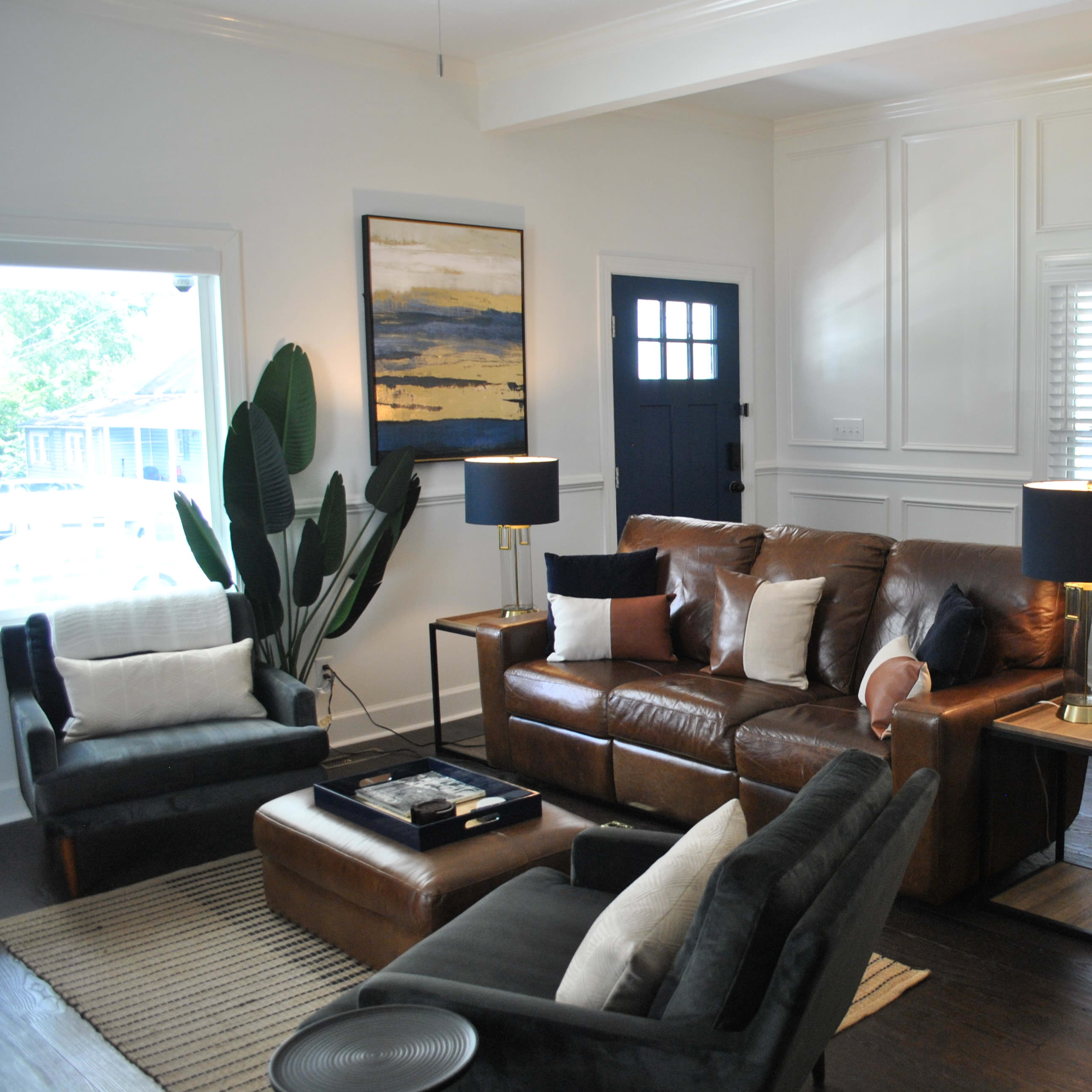 The living room features a brown leather sofa, two dark chairs, a wooden side table, and decorative plants against a backdrop of white shiplap walls and large windows.