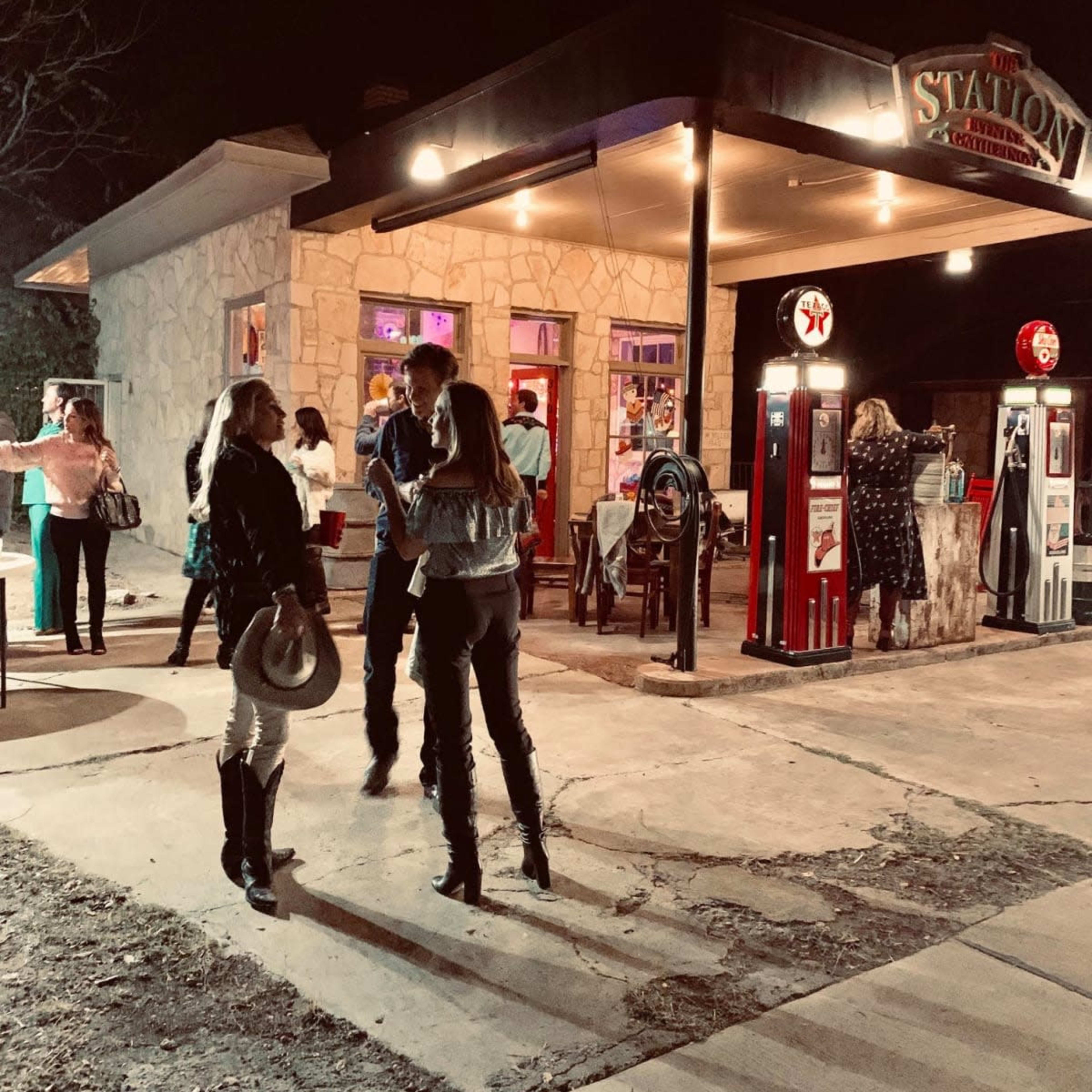 A group of people mingles outside a stone building with neon lights and vintage gas pumps on a busy street at night.