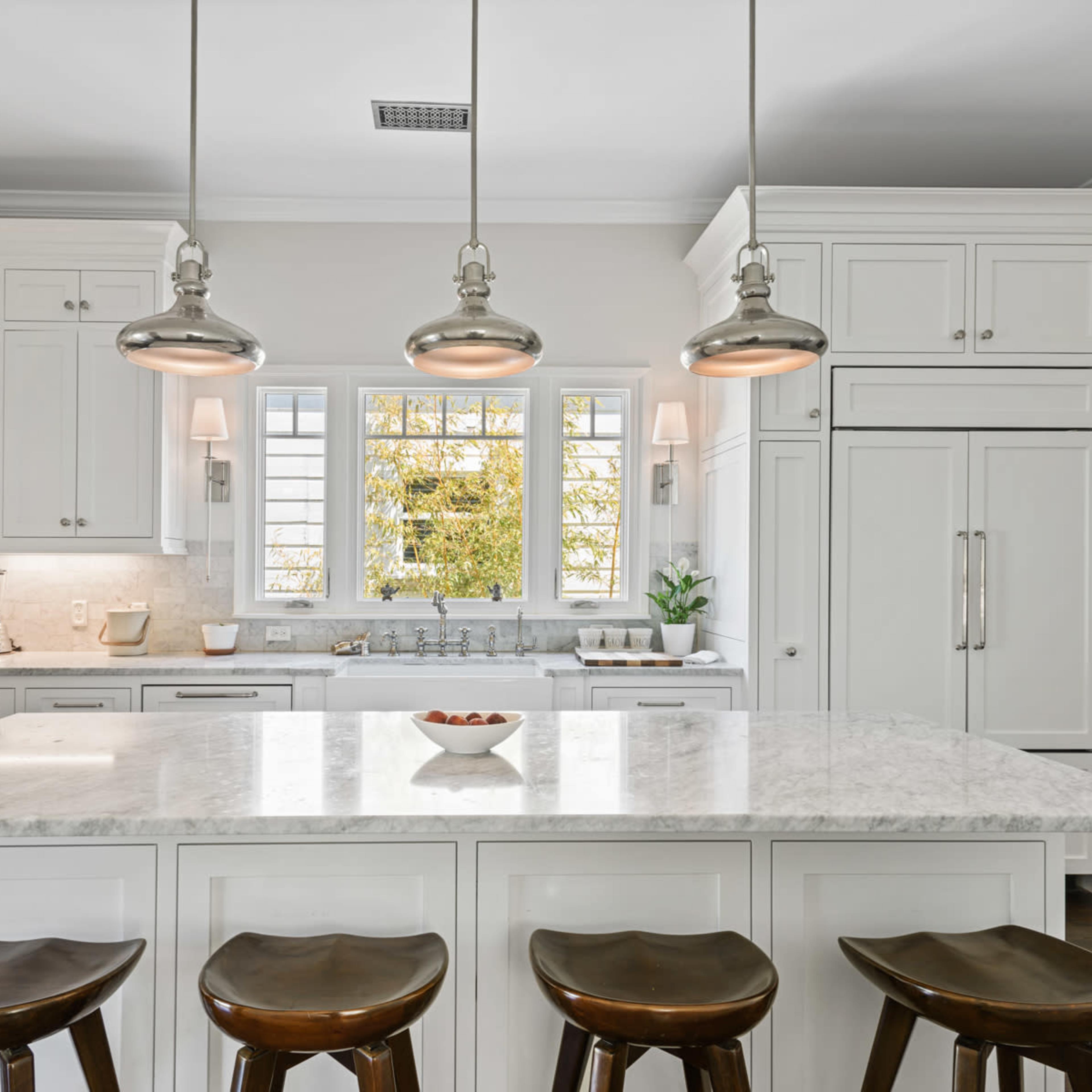 A modern kitchen features white cabinetry, a large marble island with four wooden stools, and three pendant lights hanging above.