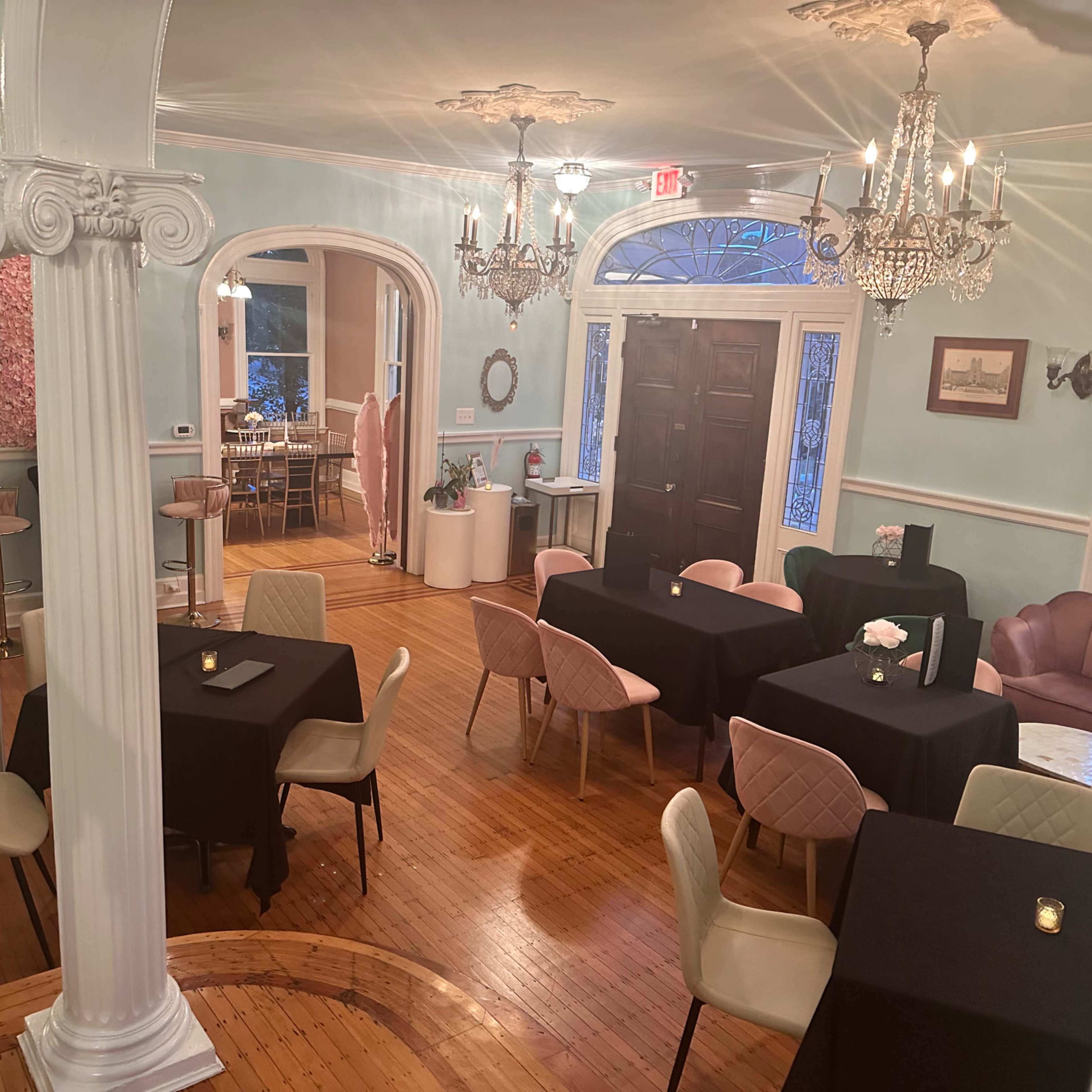 A dining area with several tables set for guests, featuring pale blue walls and decorative columns, illuminated by chandeliers.