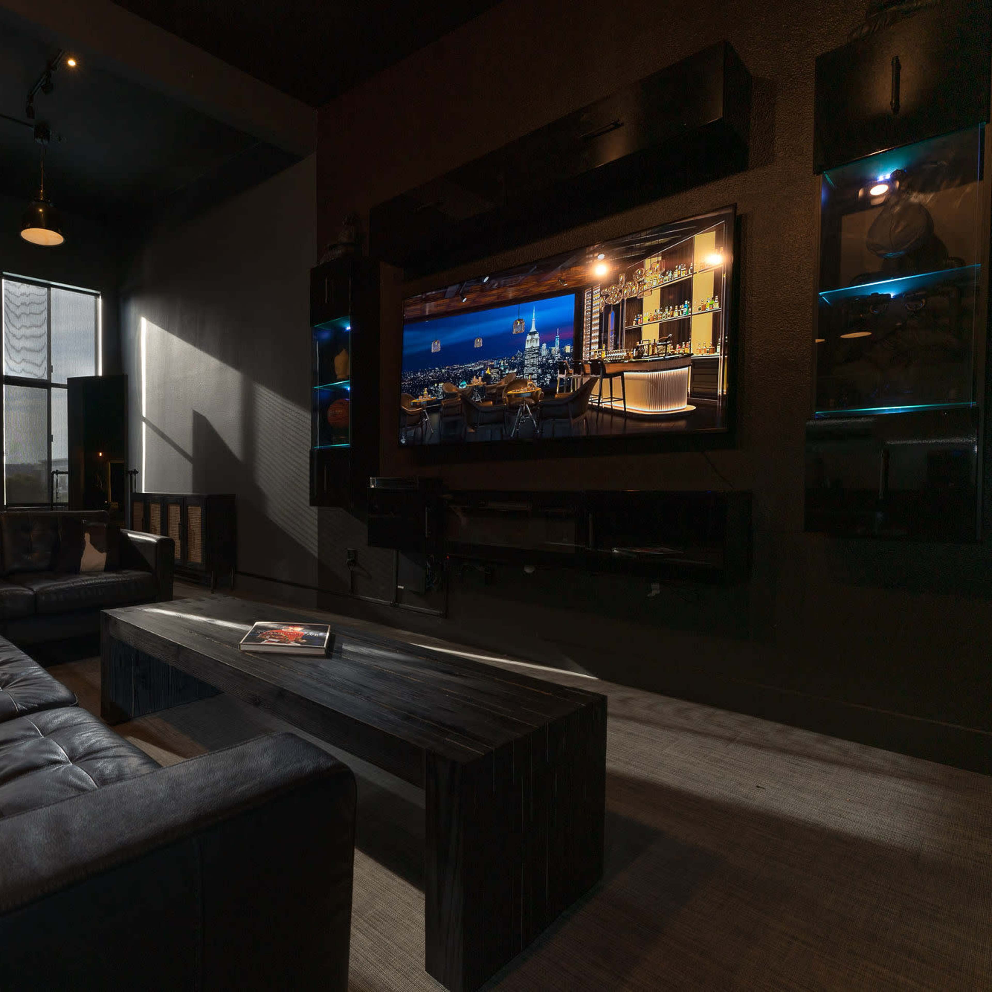 A modern living room with a black leather sofa, a wooden coffee table, and a large flat-screen TV mounted on the wall, illuminated by natural light from the windows.