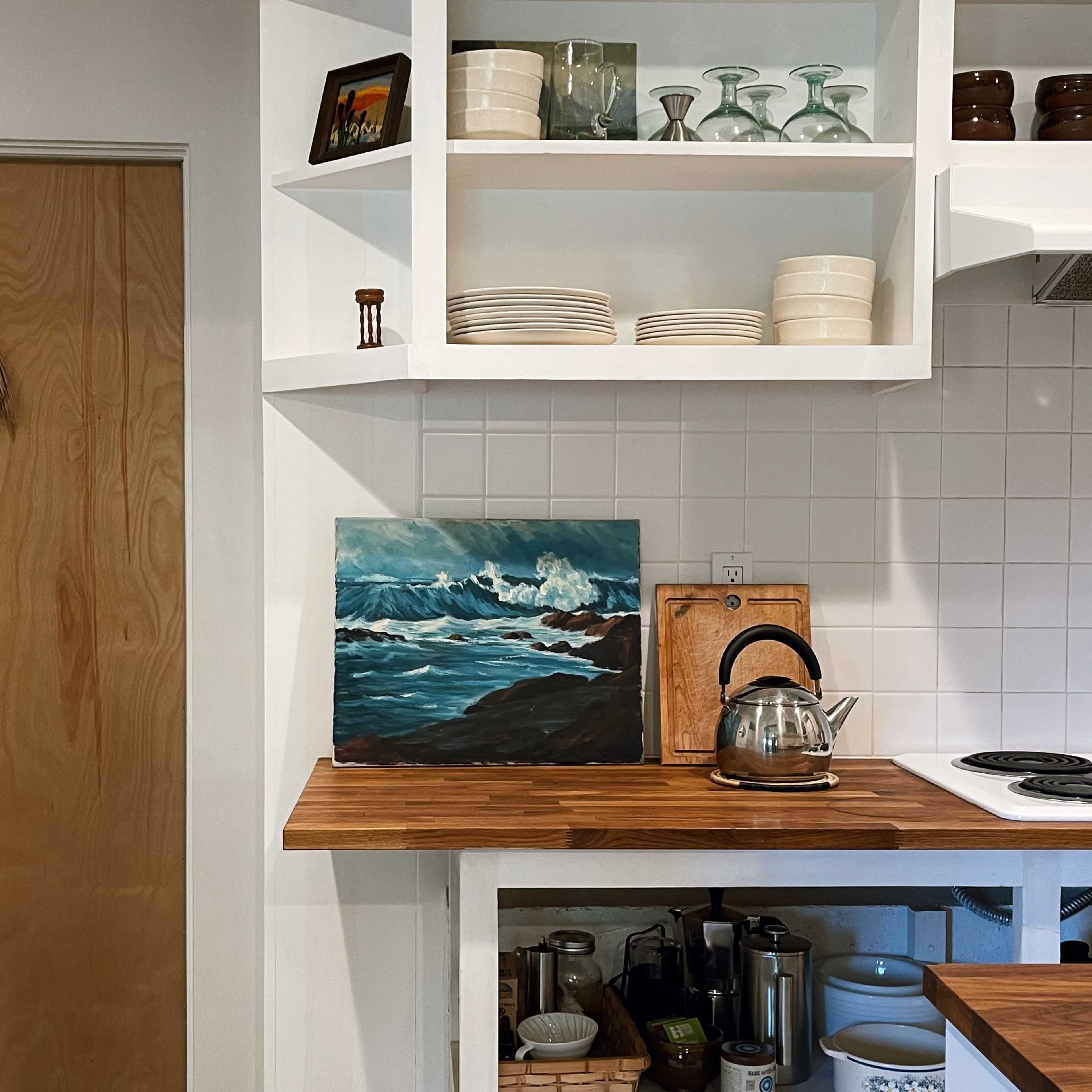 A kitchen scene features a wooden countertop with a kettle and an artwork, alongside shelves displaying glassware and dinnerware, next to a closed door.