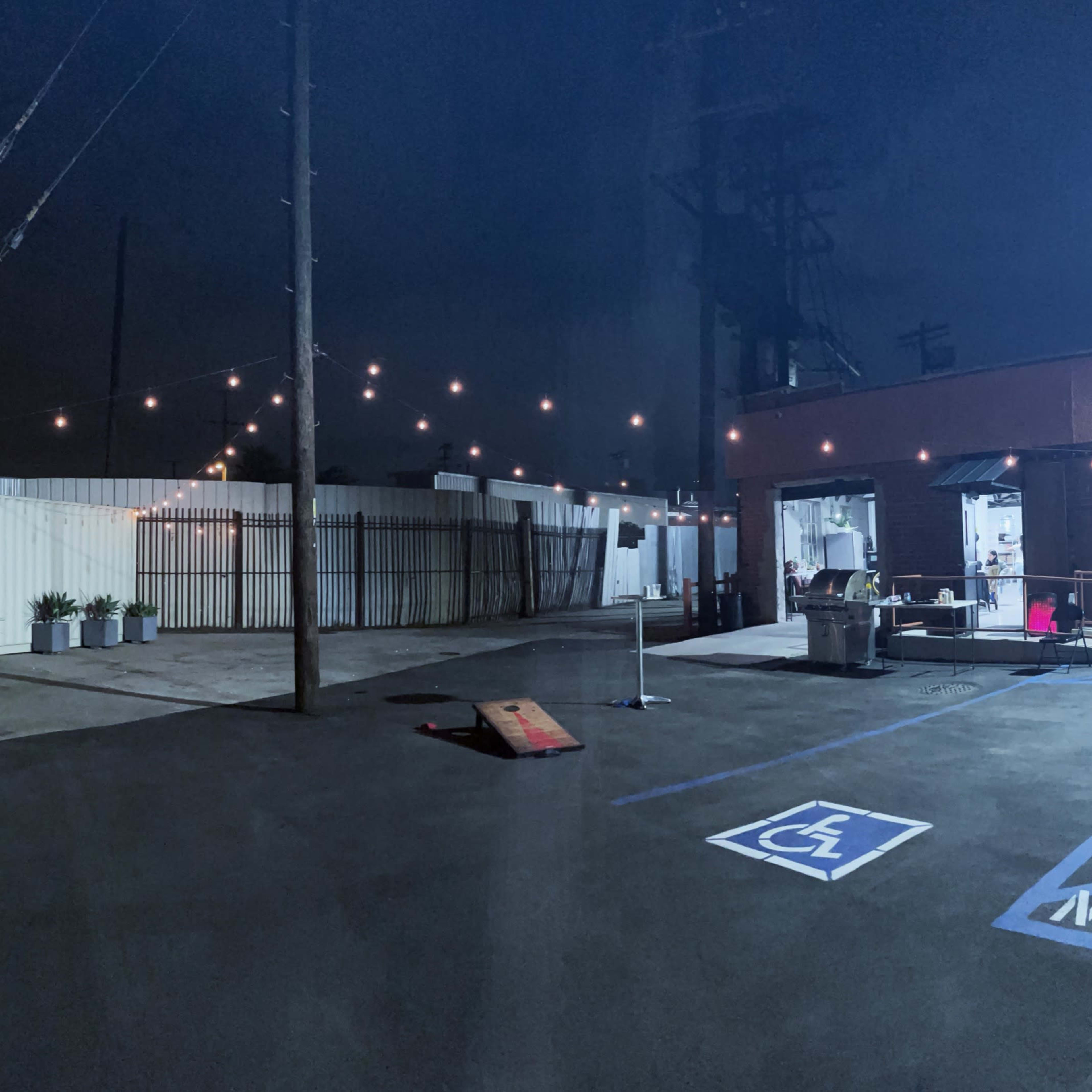 A dimly lit outdoor area adjacent to a building, featuring string lights overhead, a marked wheelchair-accessible parking space, and a few potted plants lining the perimeter.