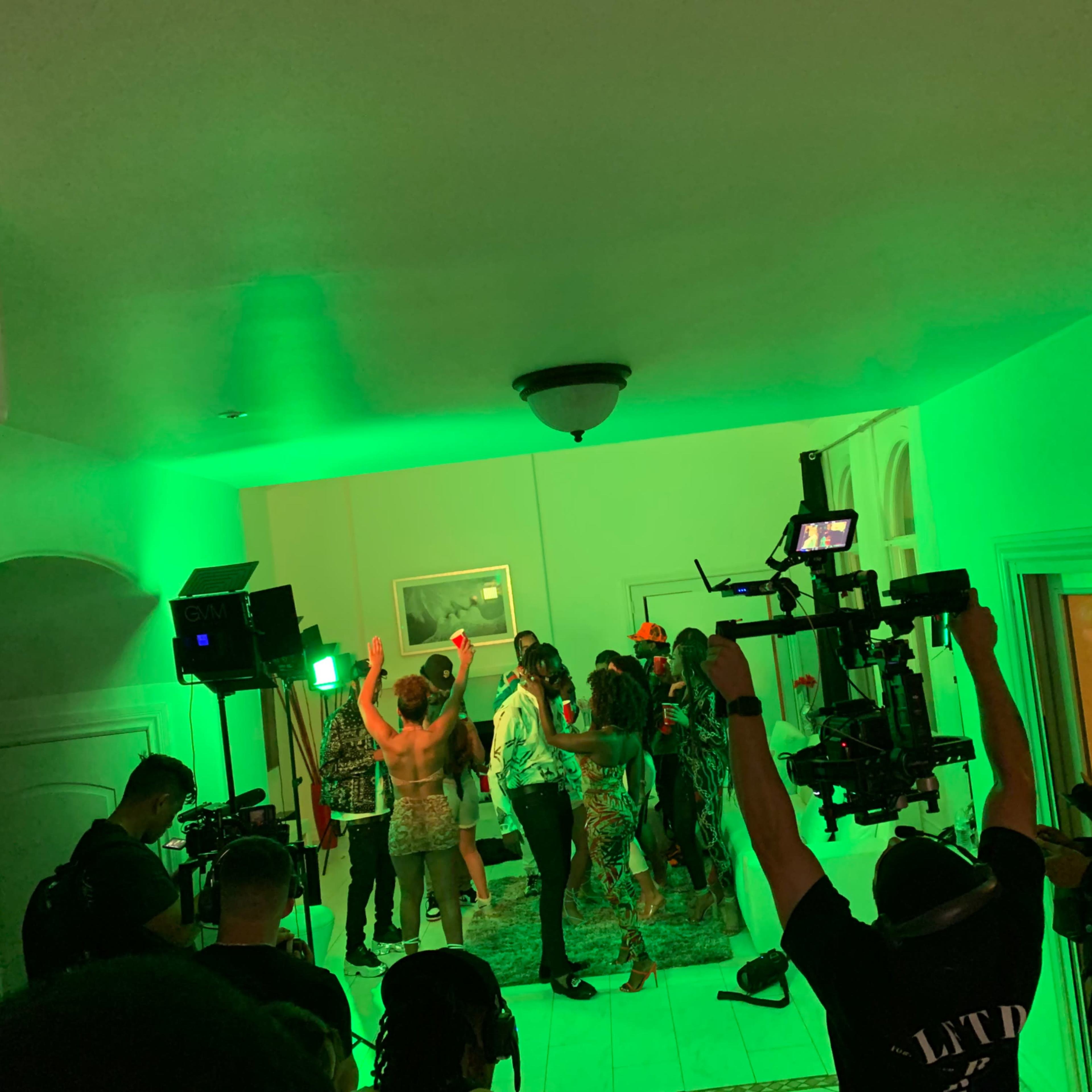 A group of people dances in a brightly lit room while crew members operate cameras and lighting equipment.