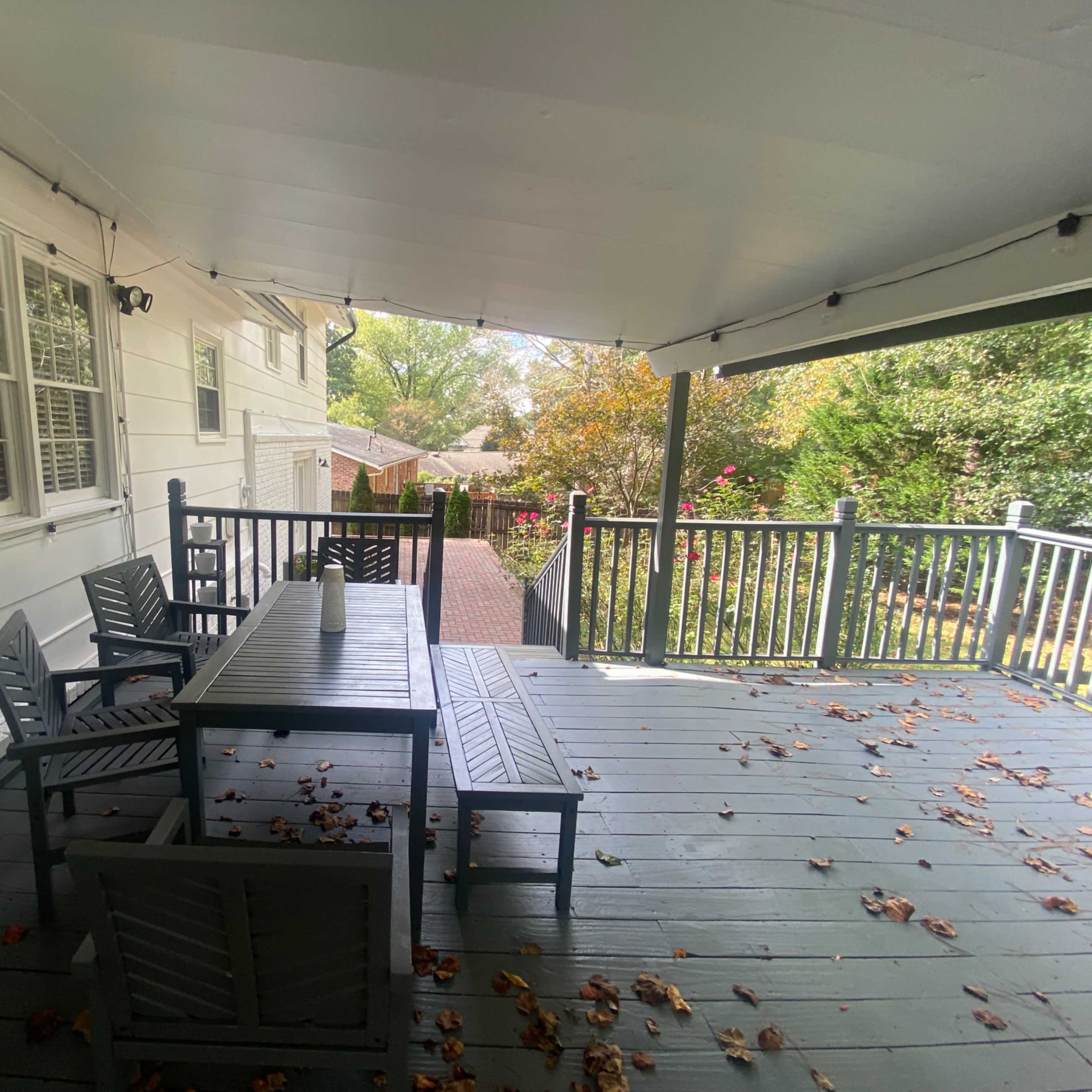 A covered deck features a wooden table and chairs surrounded by fallen leaves, with a view of a yard and garden beyond.