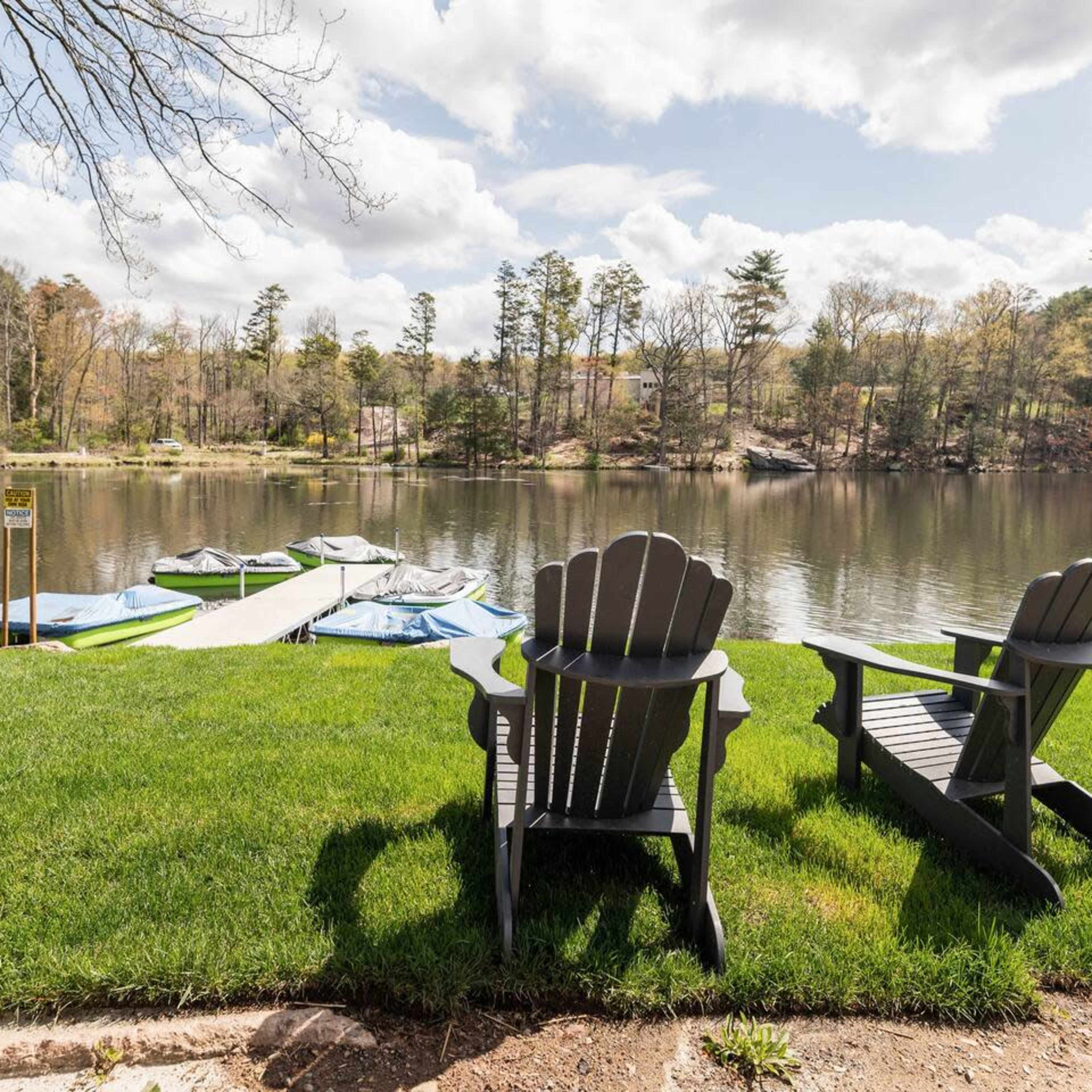 Two black adirondack chairs sit on green grass beside a calm lake, with a boat dock and an American flag in the background.