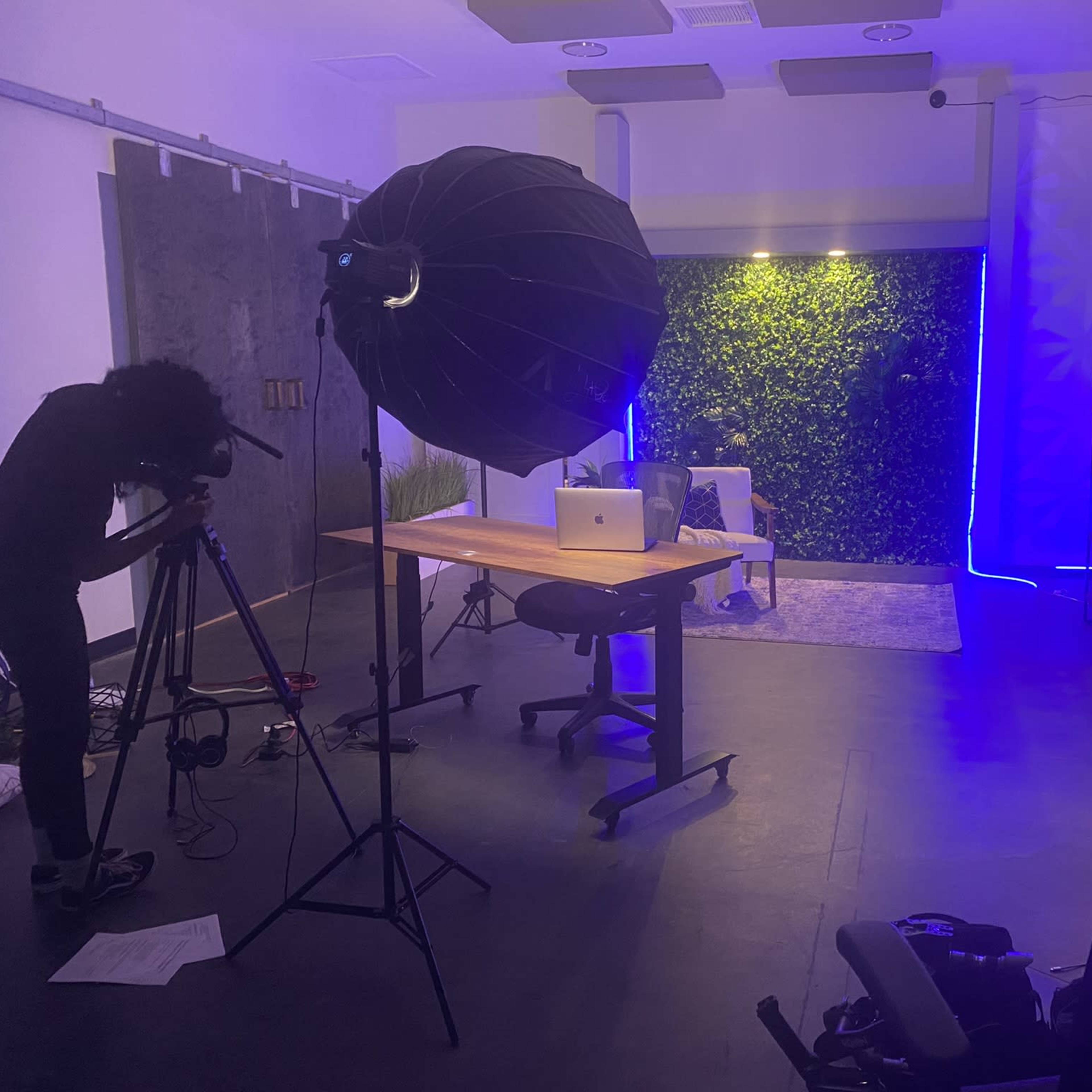 A photographer sets up a camera and lighting in a modern studio with a desk, laptop, and a greenery backdrop.
