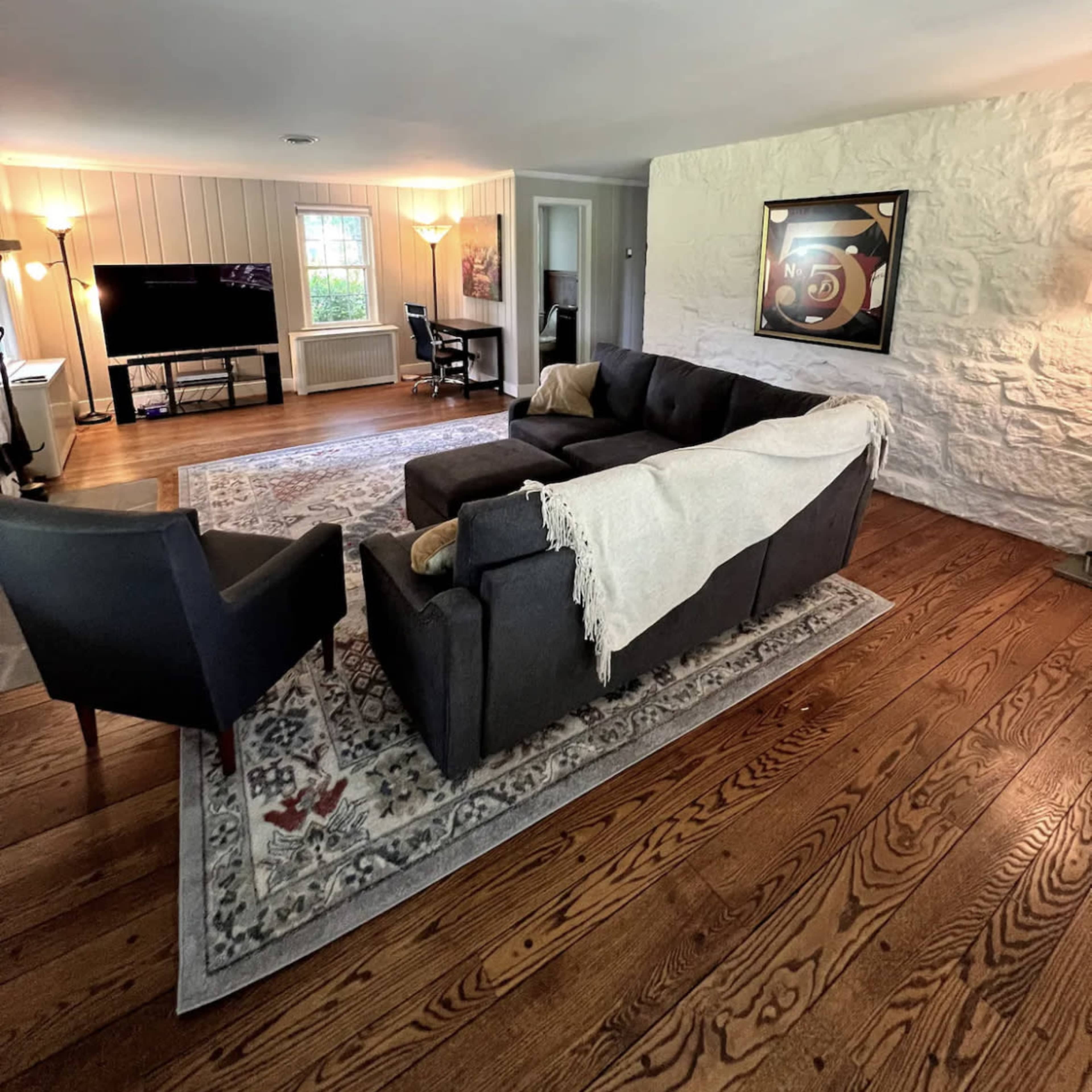 A living room with a dark sofa and armchair positioned on a wooden floor, a large area rug, a television, and a stone wall.