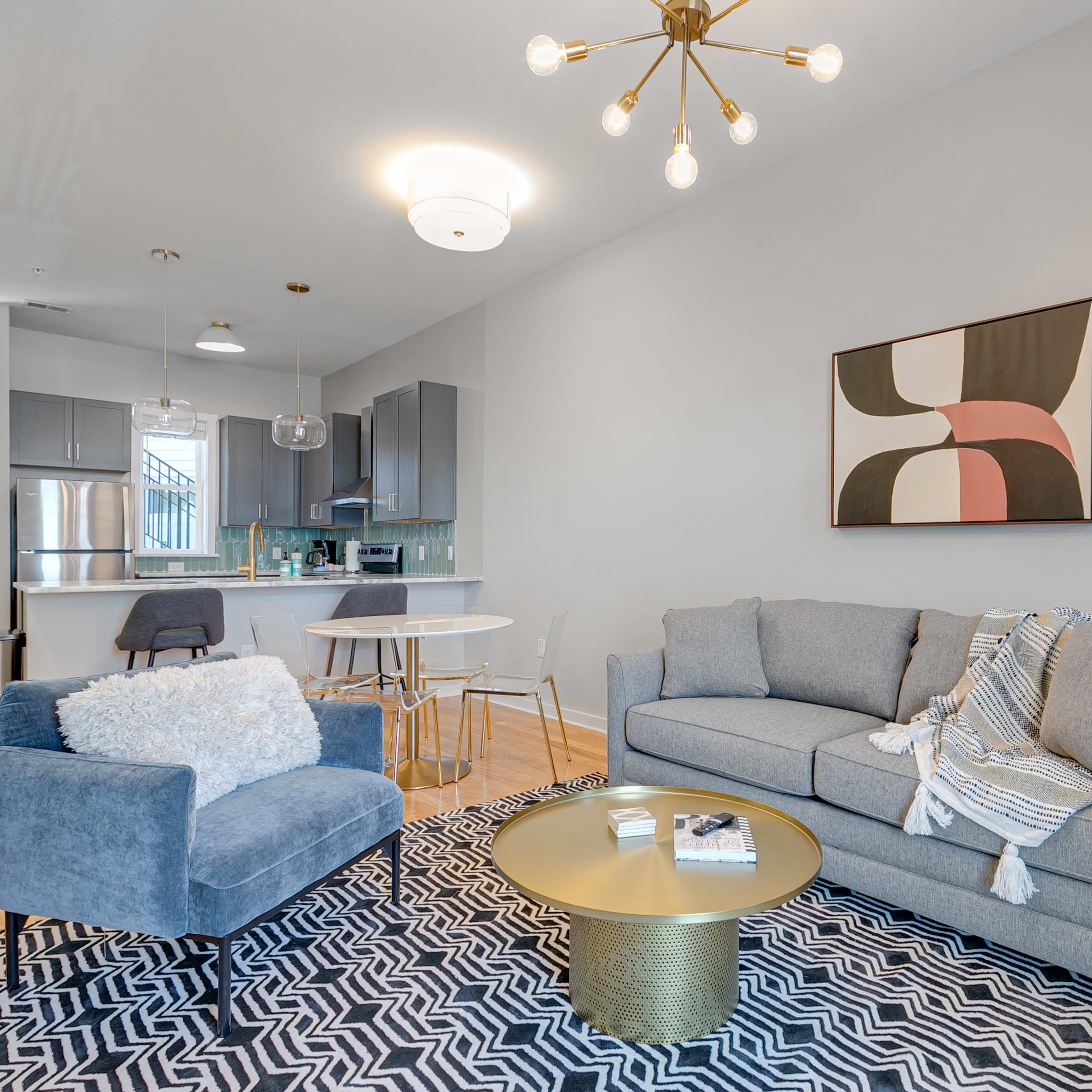 A modern living space featuring a gray couch, a blue accent chair, a round gold coffee table, and a dining area with a small white table and chairs.