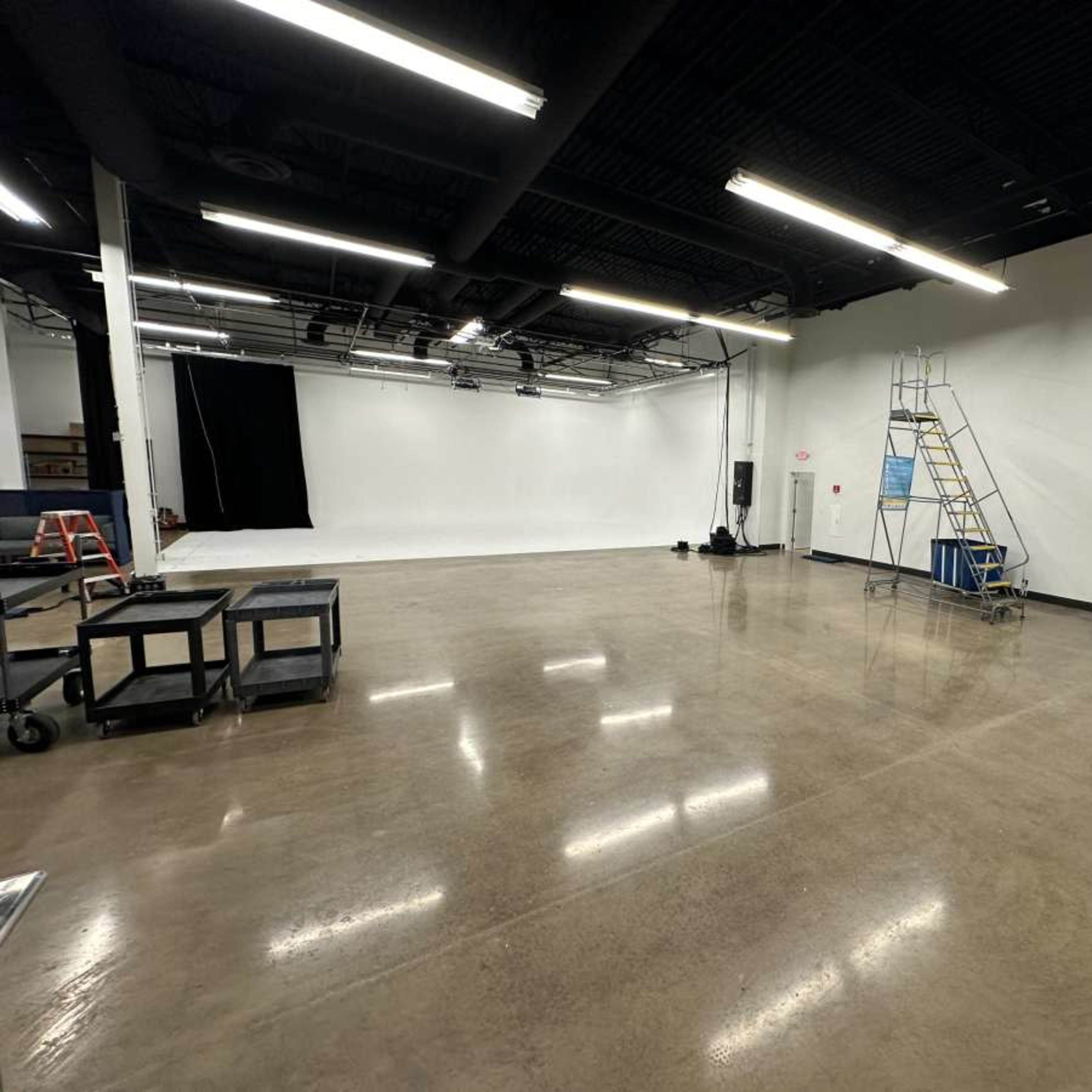 A spacious, empty studio with smooth concrete flooring, black drapery on one wall, and a tall ladder near the center.