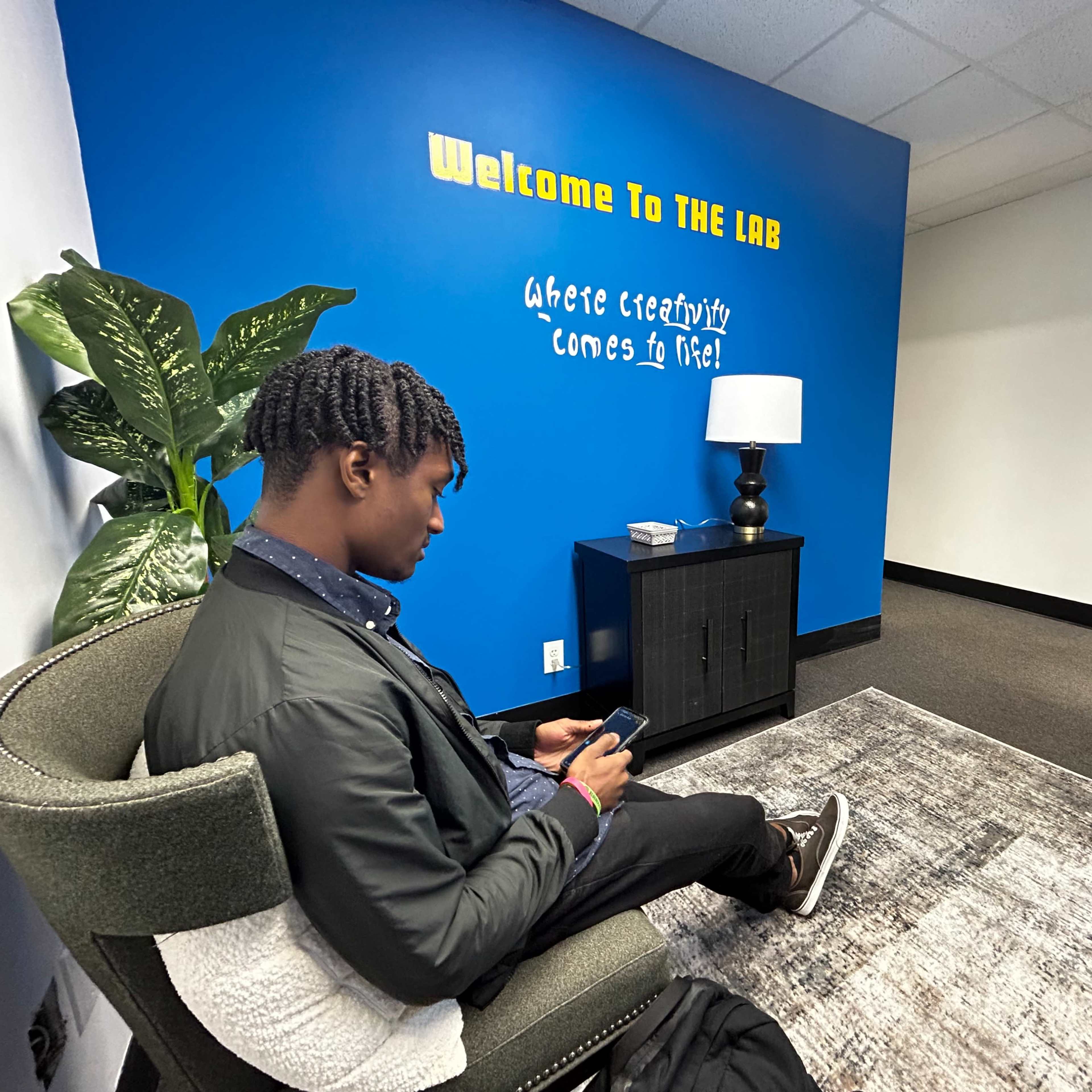 A young man sits on a chair while using his phone in a room with a blue wall that features the phrase "Welcome To THE LAB" and a bedside lamp.