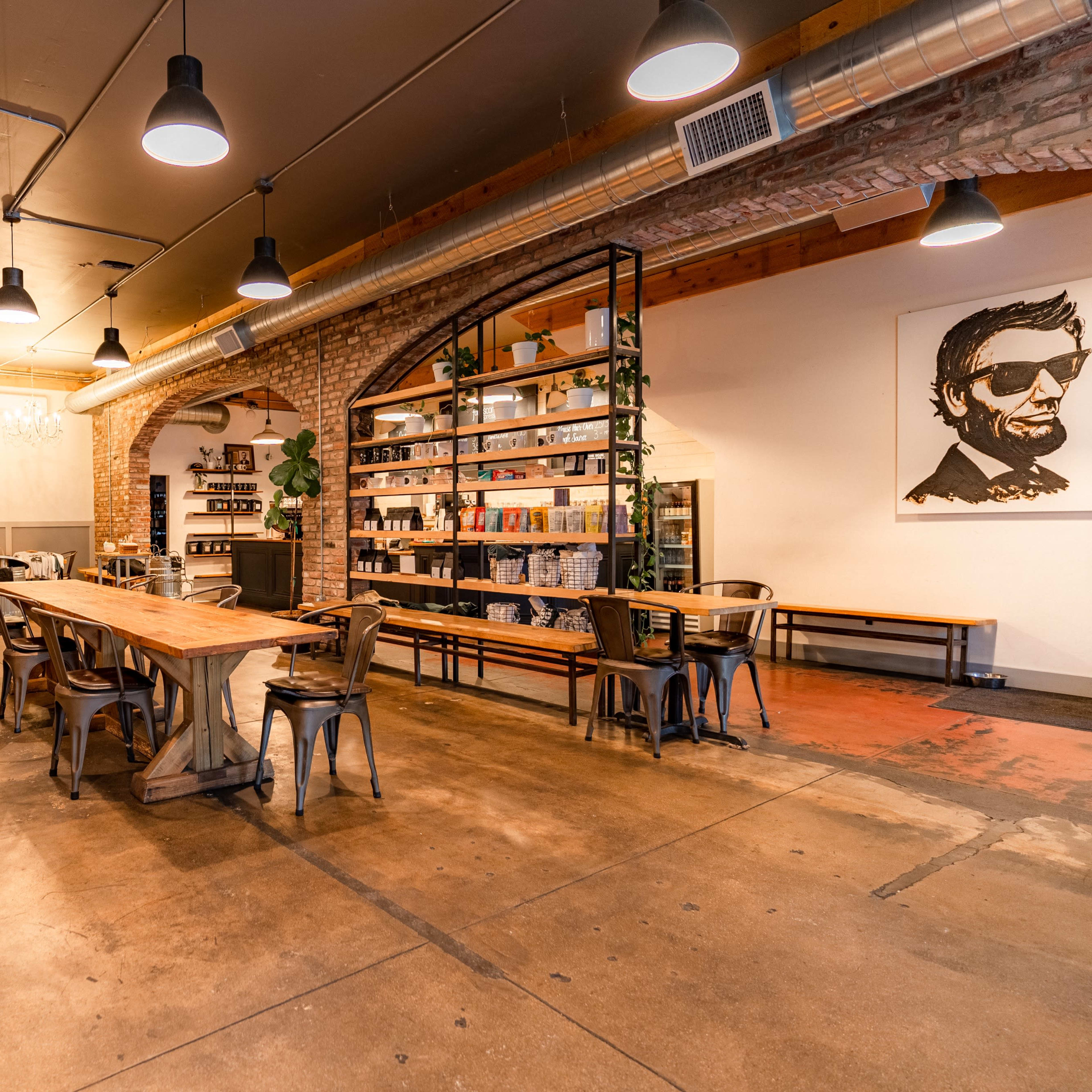 An interior of a spacious café featuring wooden tables, industrial-style lighting, a brick wall, and a shelf filled with various items.