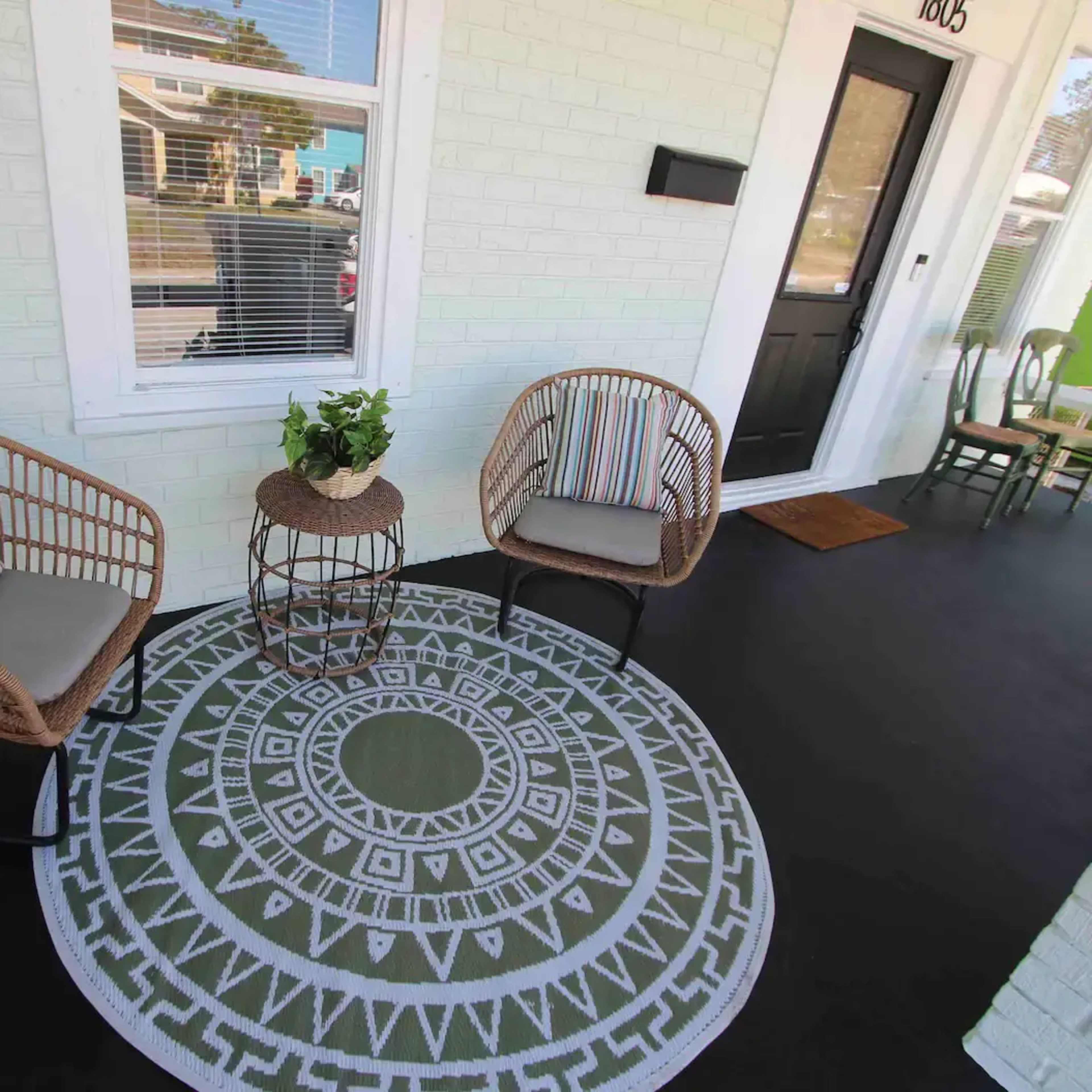 A porch area featuring two wicker chairs with cushions, a round decorative table, and a patterned outdoor rug.