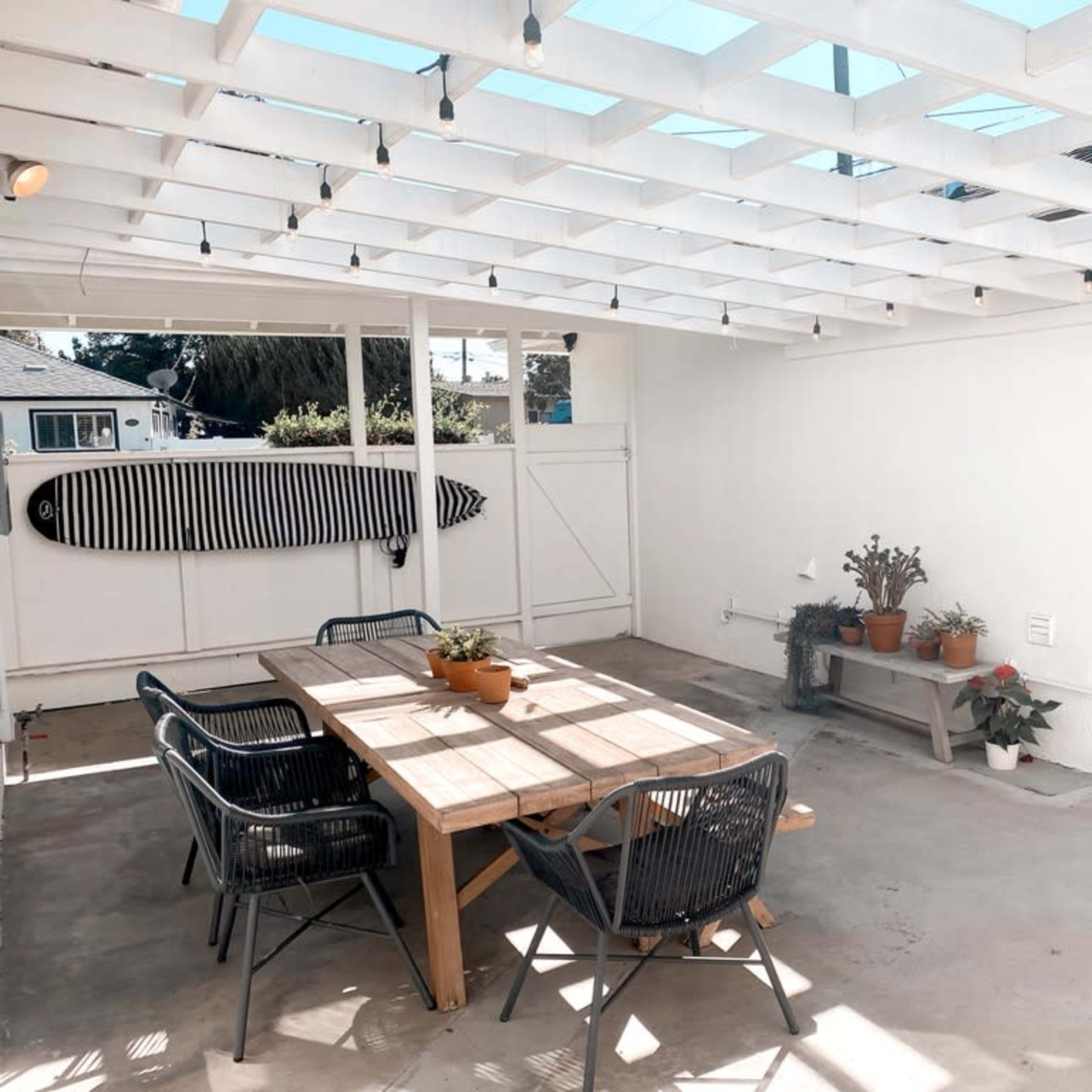 A covered outdoor patio area with a wooden table, black chairs, potted plants, and a surfboard mounted on the wall.