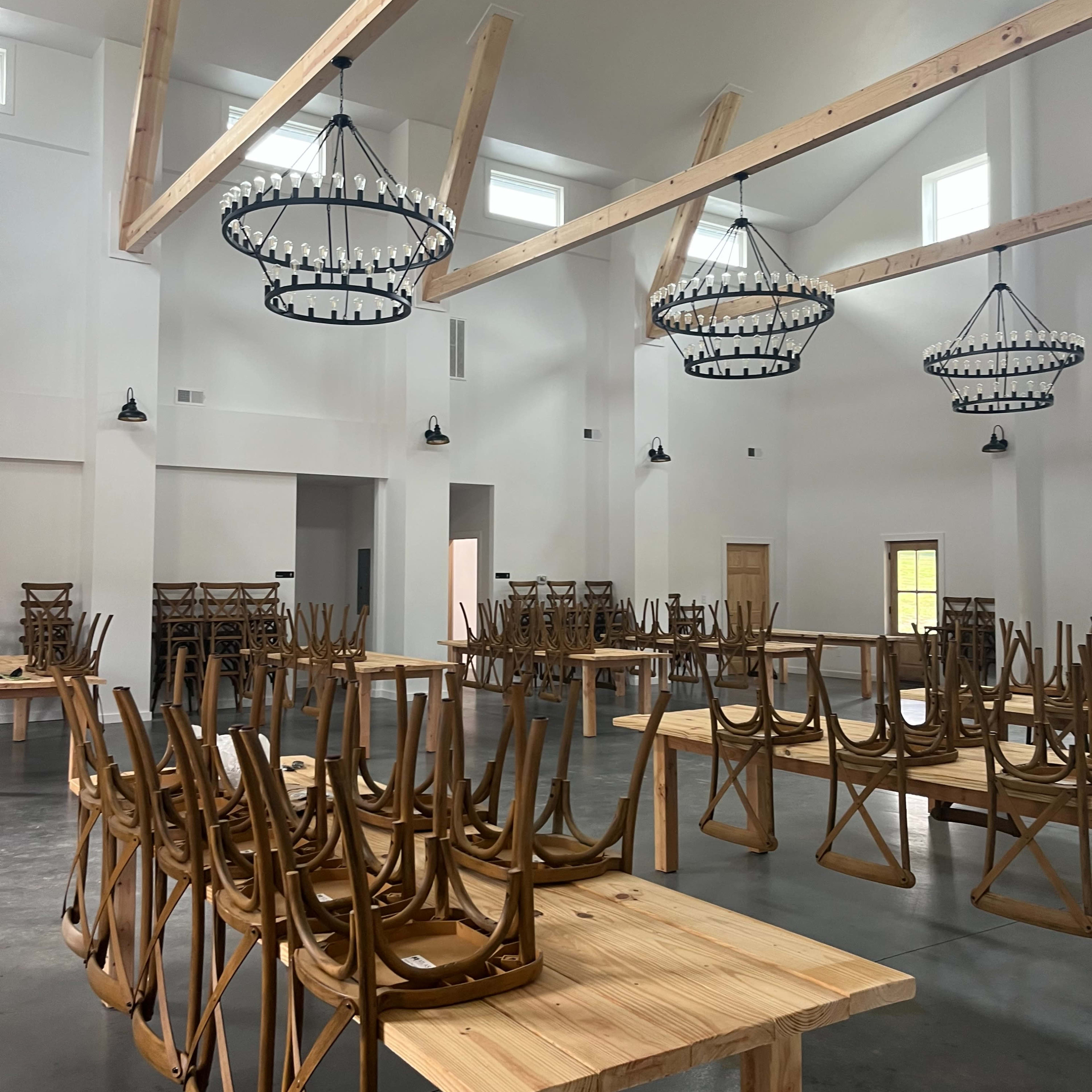 The interior of a spacious, empty room features wooden tables with chairs stacked upside down on top, and large chandeliers hanging from the ceiling.