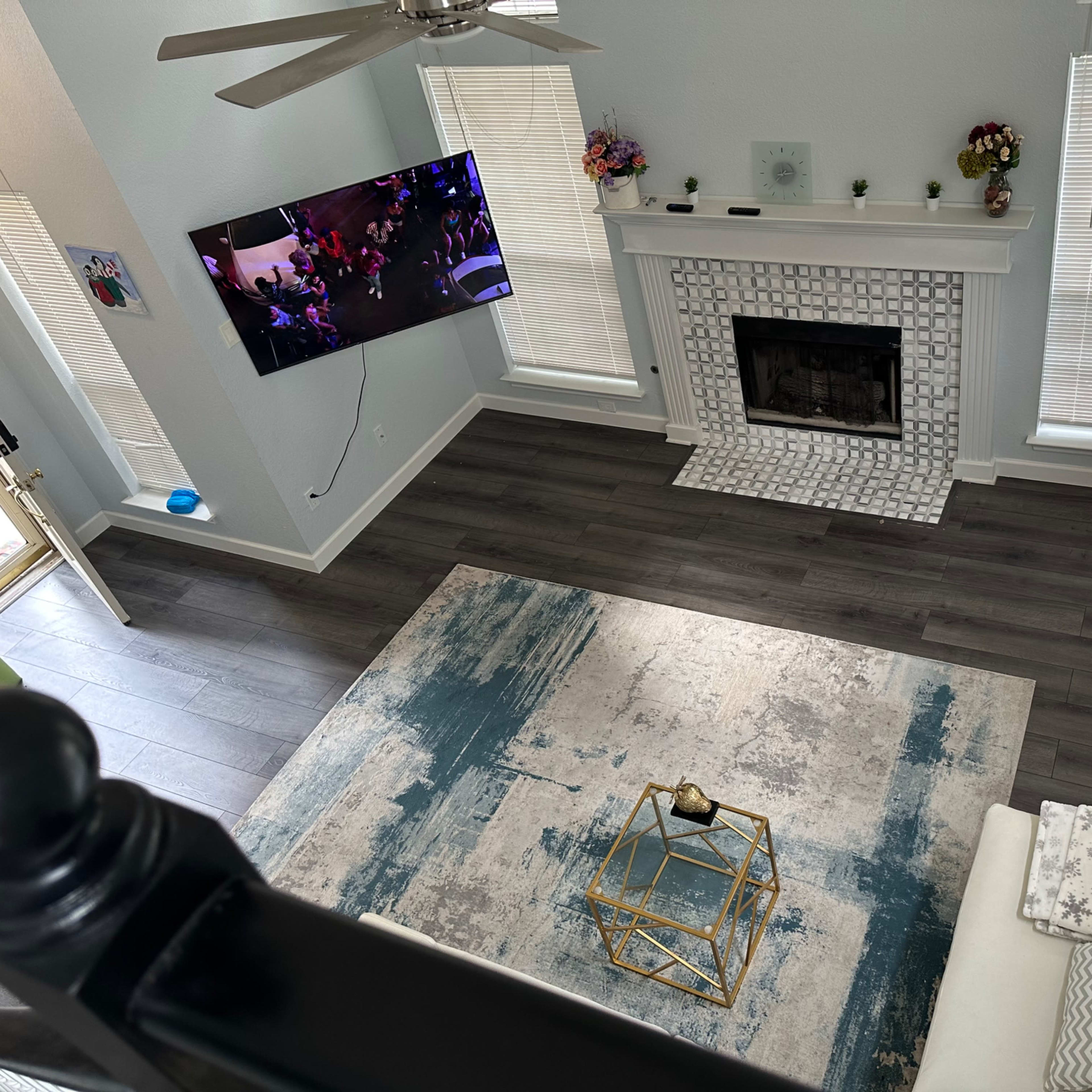 A living room with a television mounted on the wall, a fireplace with a tiled surround, a large area rug on the floor, and a light-colored sofa.