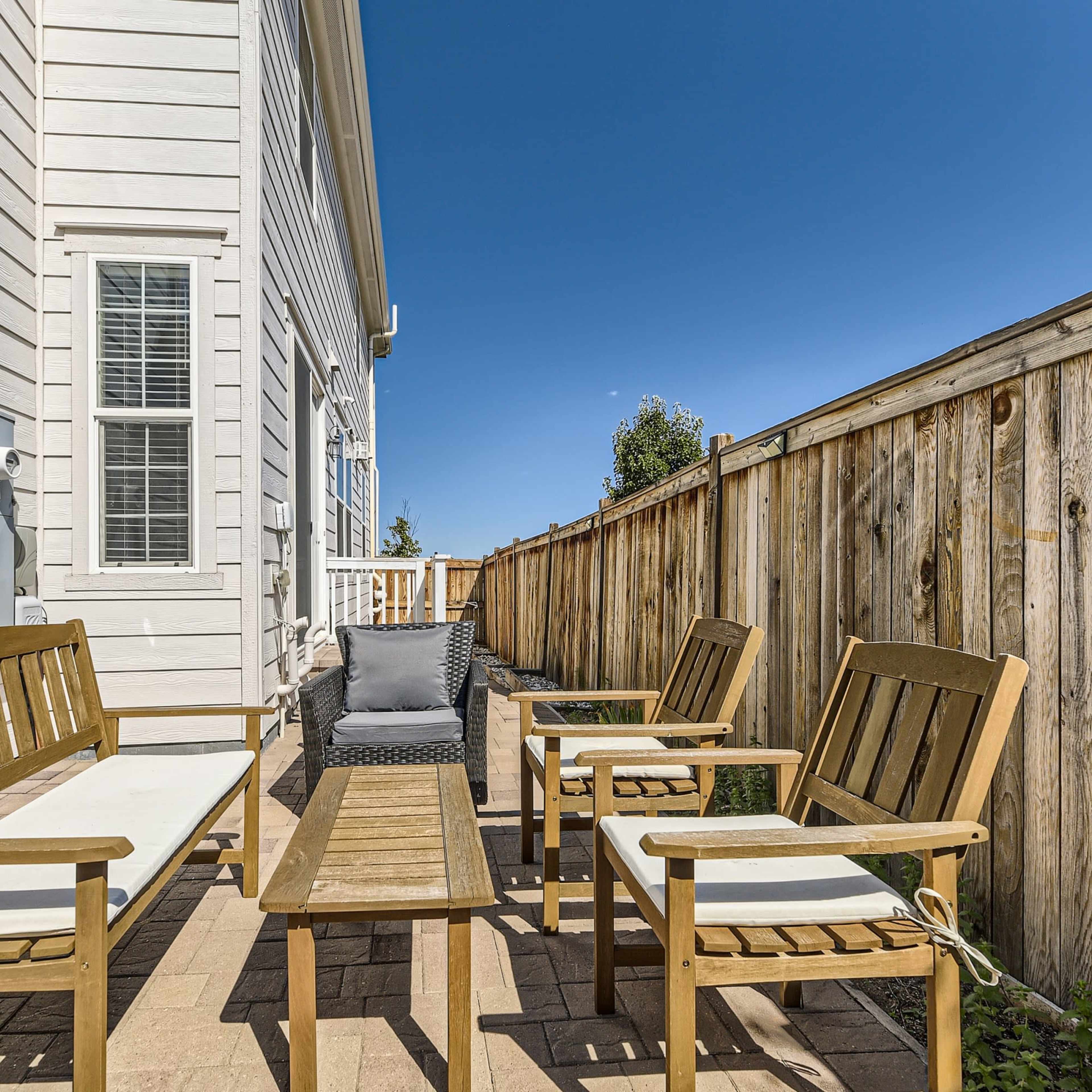 A small patio area with wooden furniture, including chairs and a table, alongside a fence and the exterior of a house.