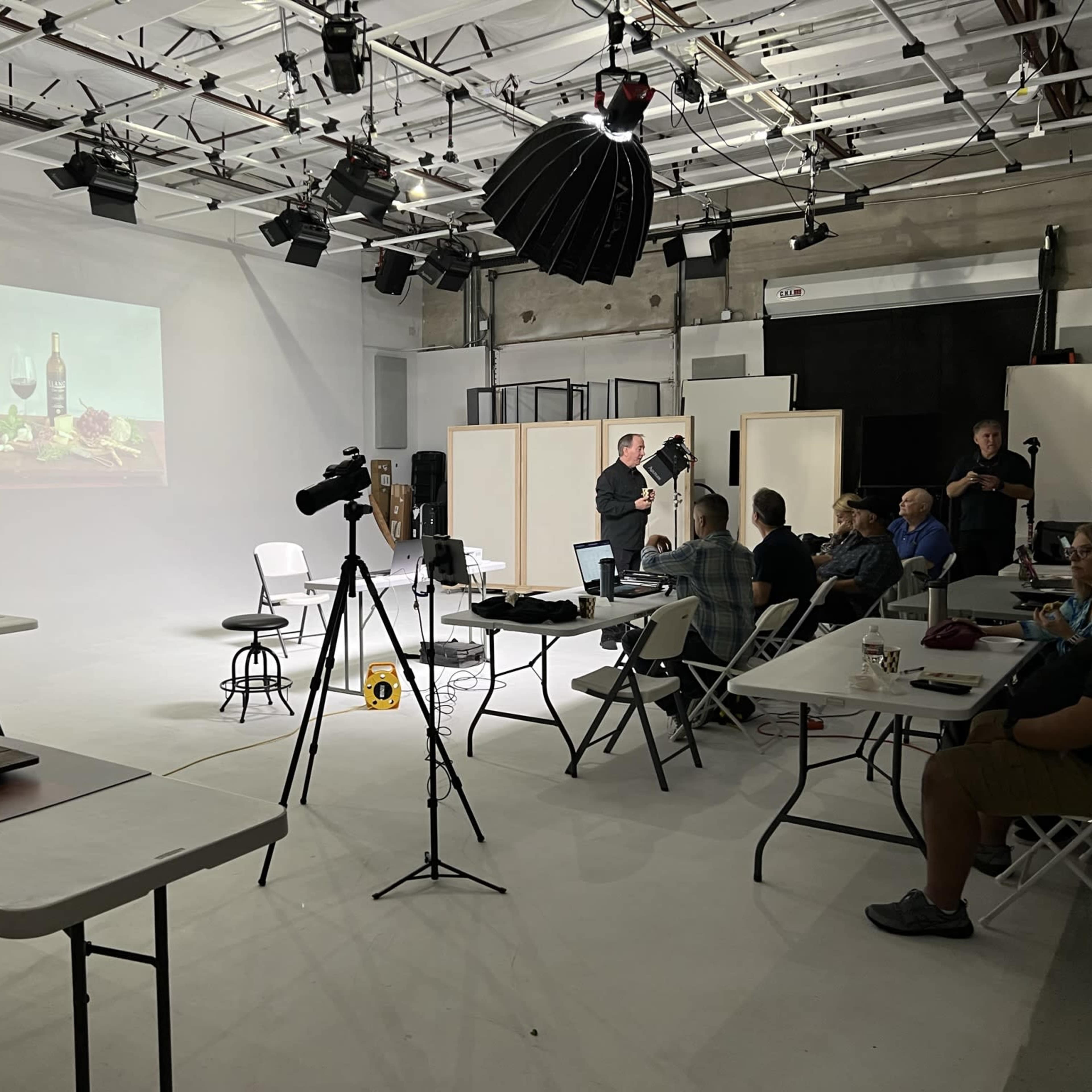 A group of people is gathered in a studio for a photography workshop, with a presentation on food and wine set up on a screen.