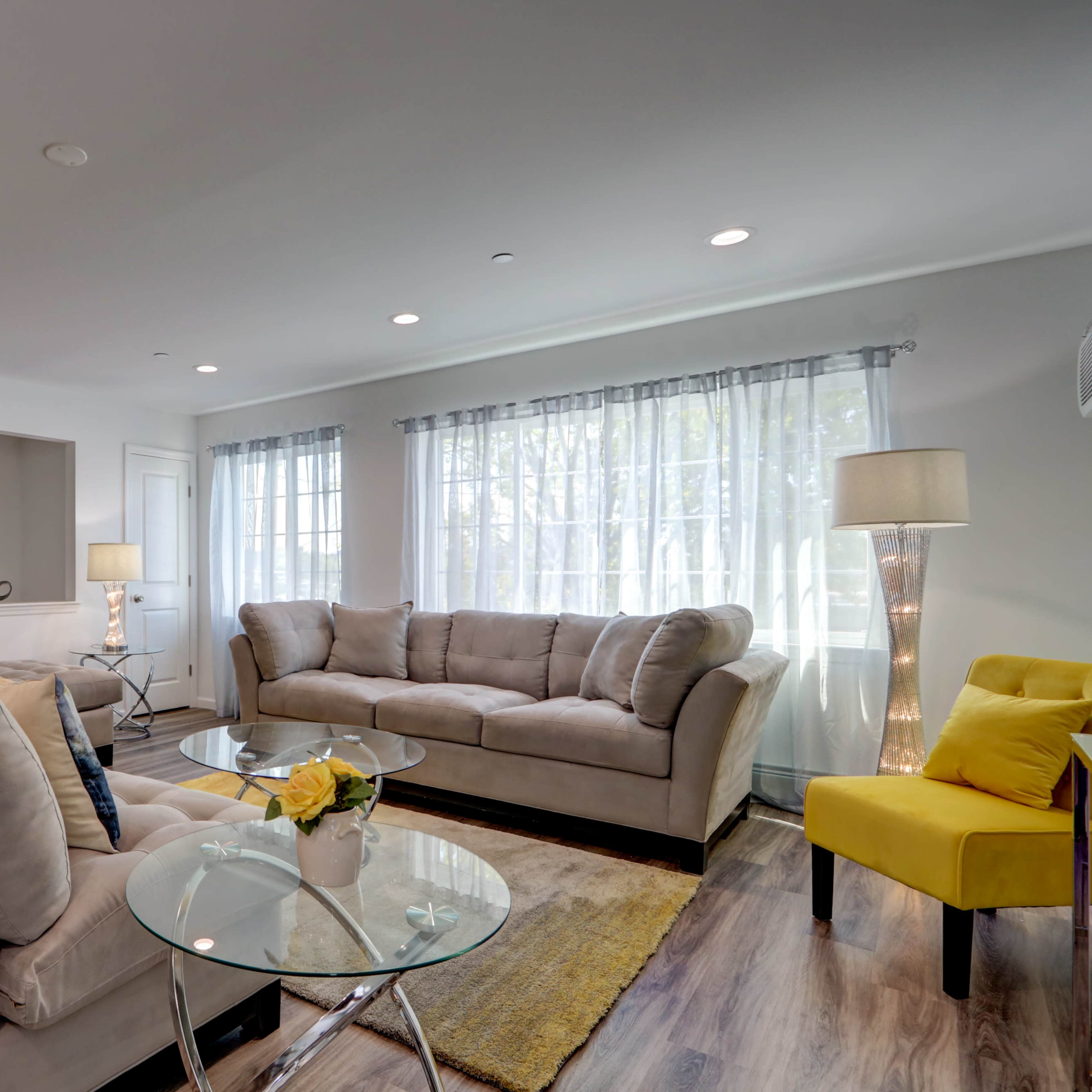 A modern living room with two beige sofas, a yellow armchair, a glass coffee table, and large windows with sheer curtains.