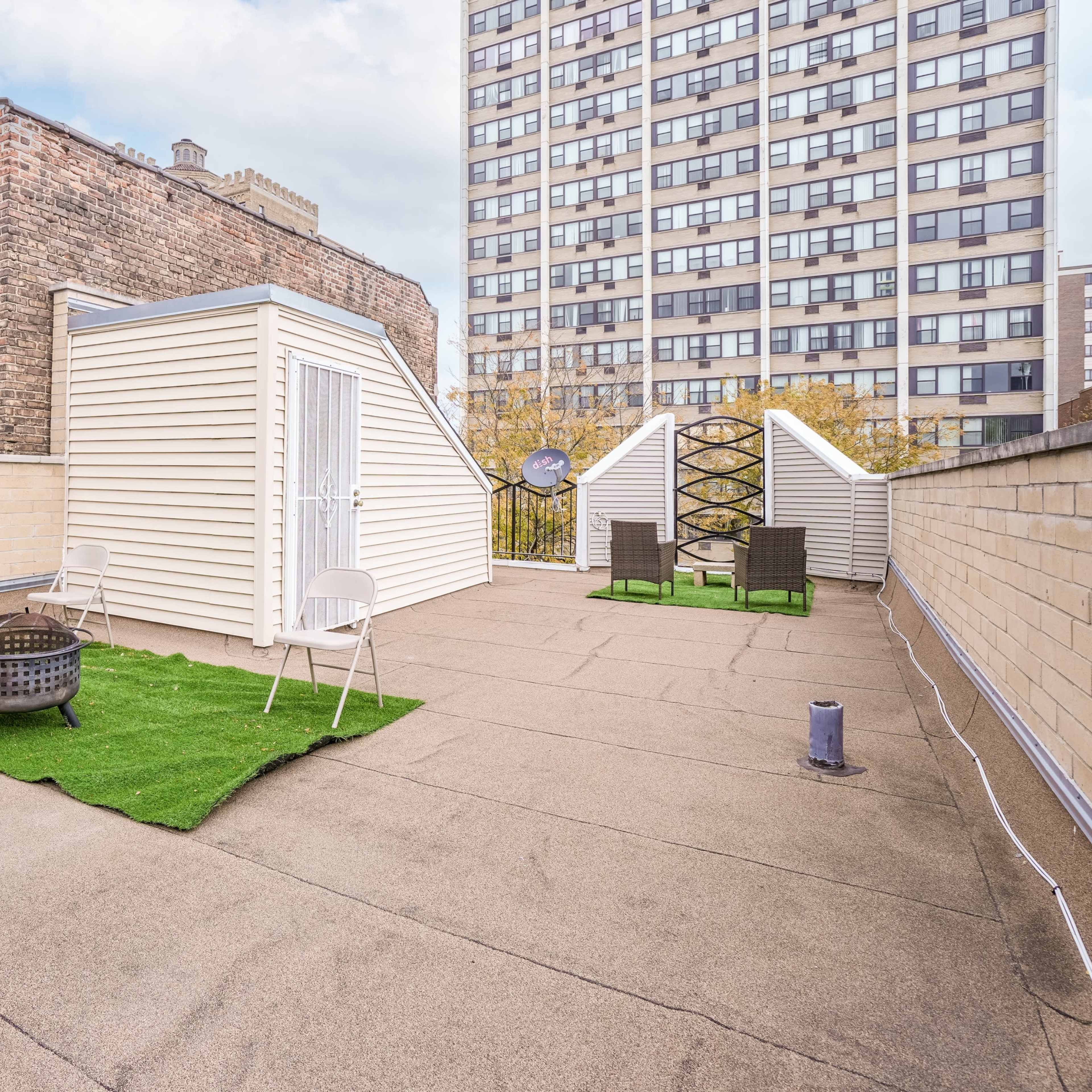 A rooftop terrace with synthetic grass, two chairs, and three small structures against a backdrop of a tall building.