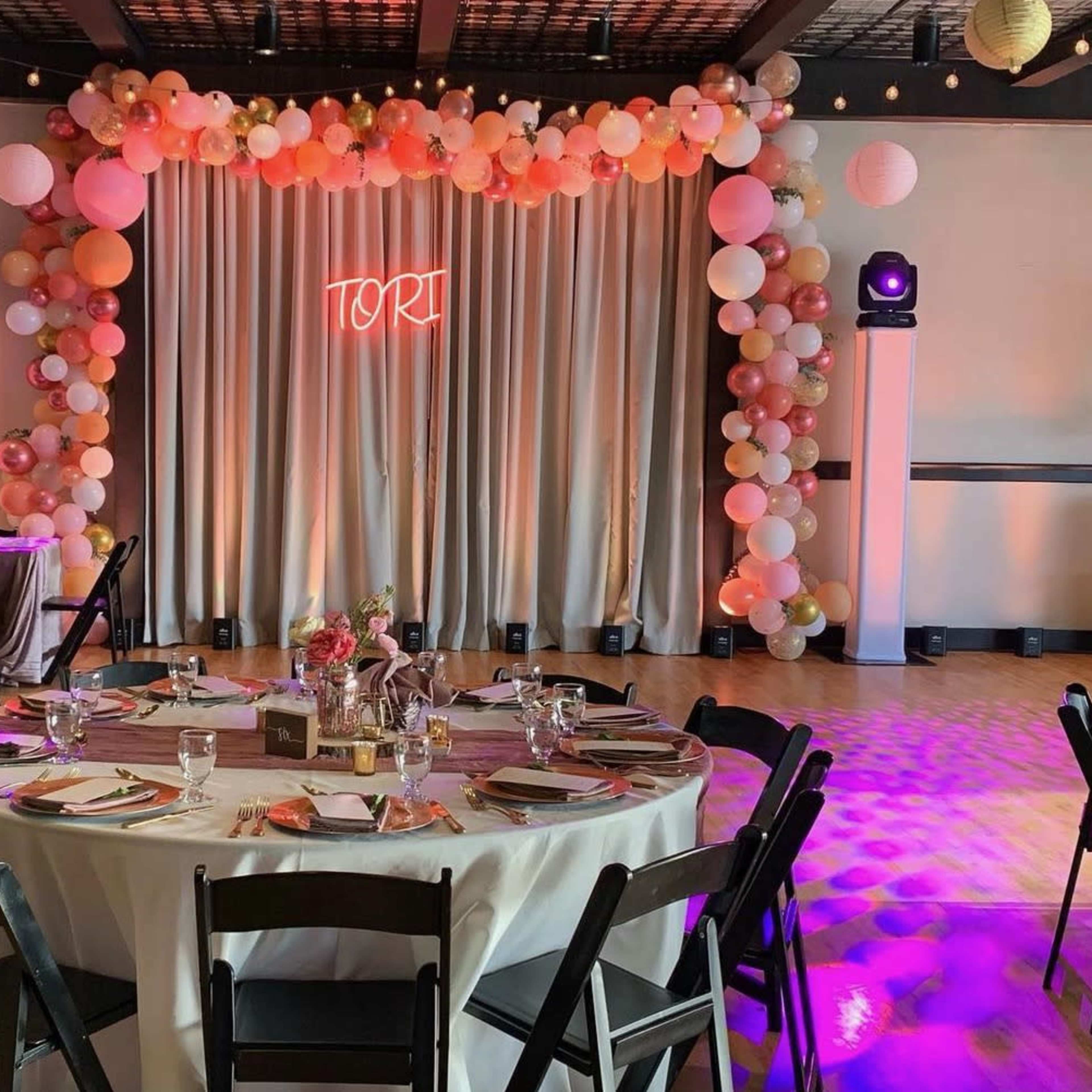 A decorated event space with a table set for dining, featuring a backdrop of balloons and a glowing sign that reads "Tori."