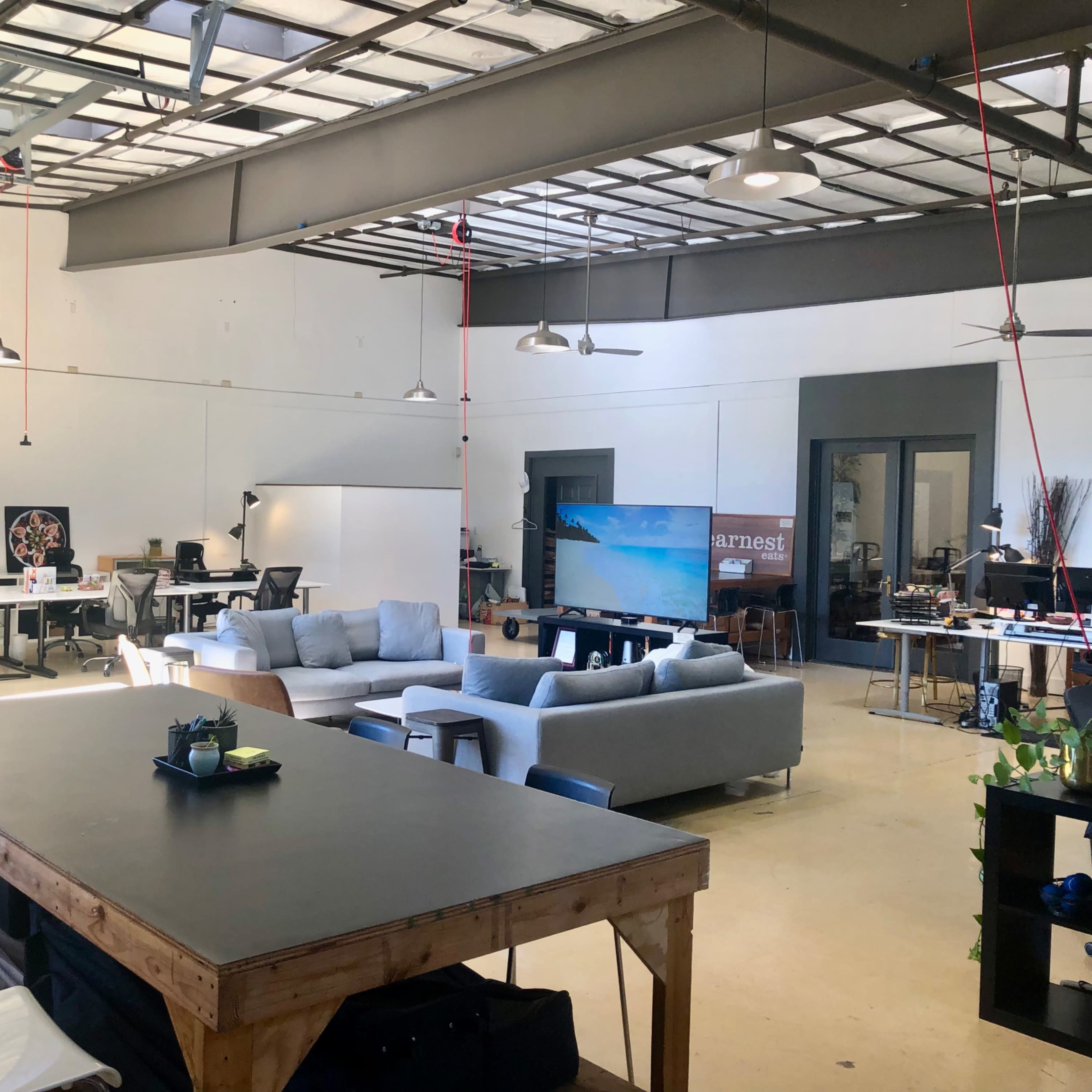 An open office space with multiple seating areas, workstations, and a large screen in a bright, minimalistic environment.