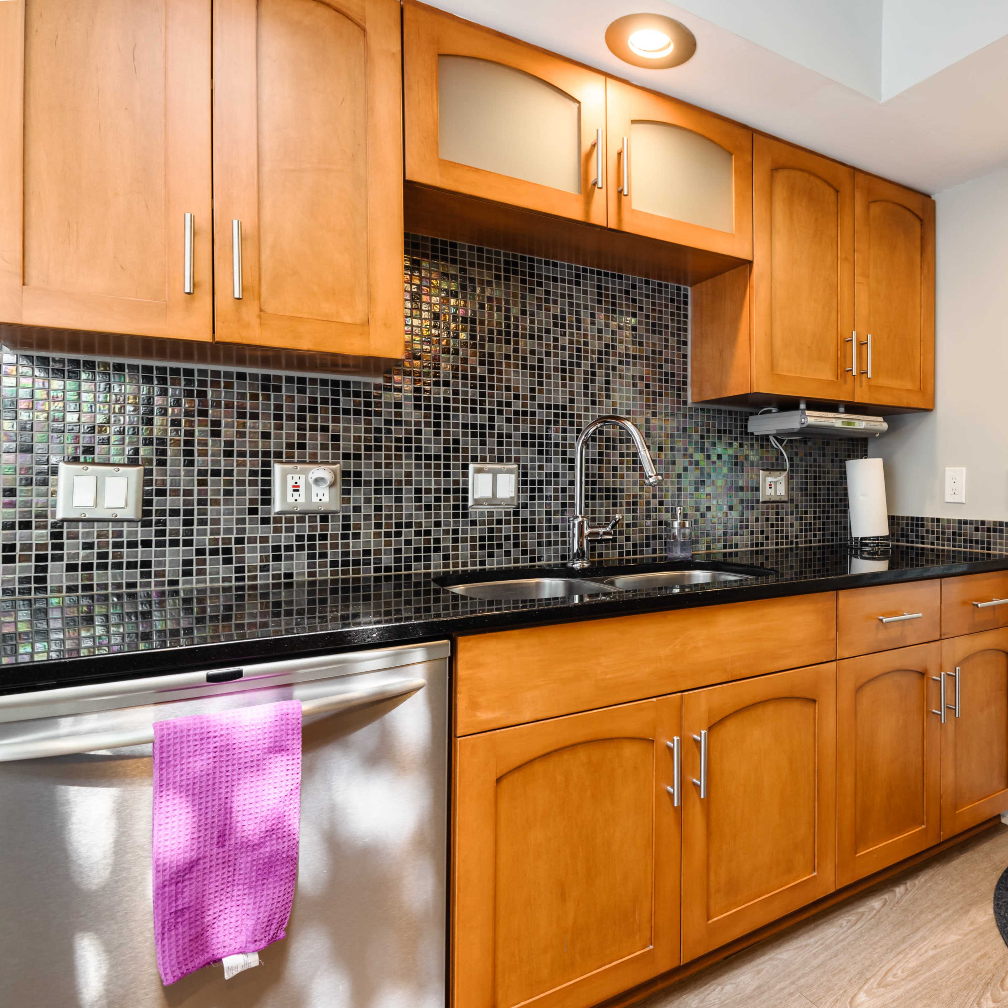 A kitchenette with wooden cabinets, a black countertop, a stainless steel dishwasher, a sink, and a glass door leading to an outdoor area.