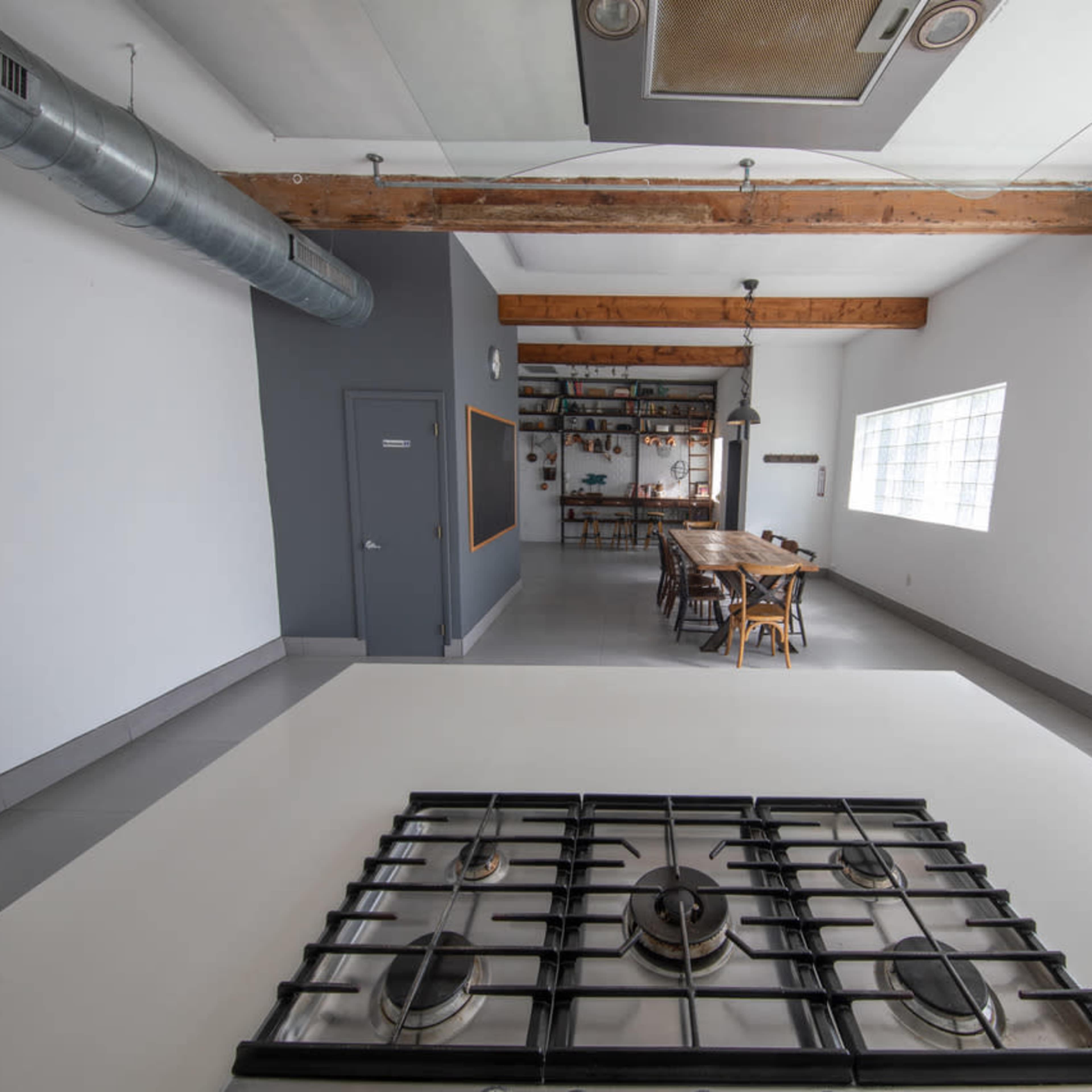 A modern kitchen with a stove in the foreground and a long dining table set in an industrial-style dining area in the background.