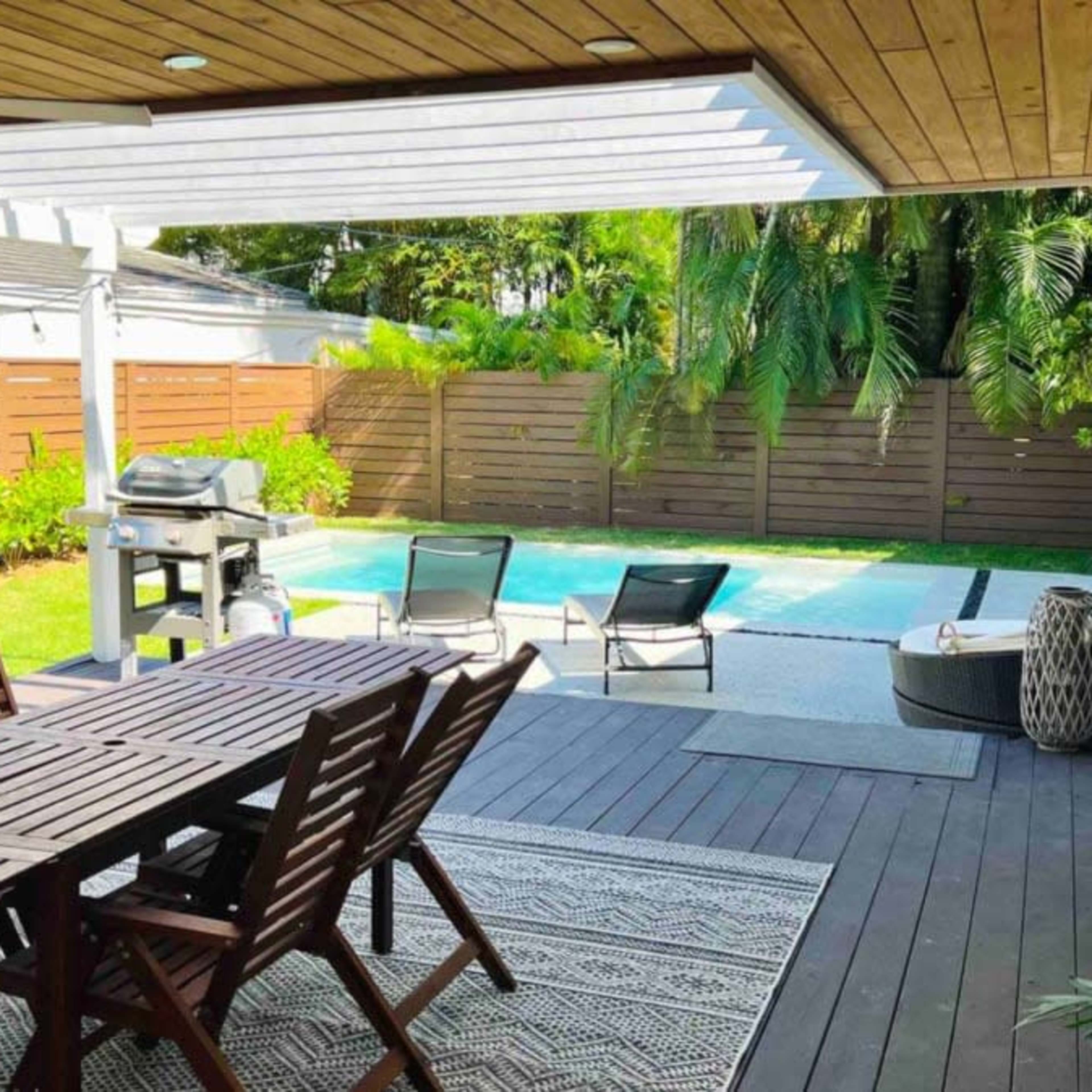 A patio area with a wooden dining table and chairs overlooking a swimming pool surrounded by greenery.