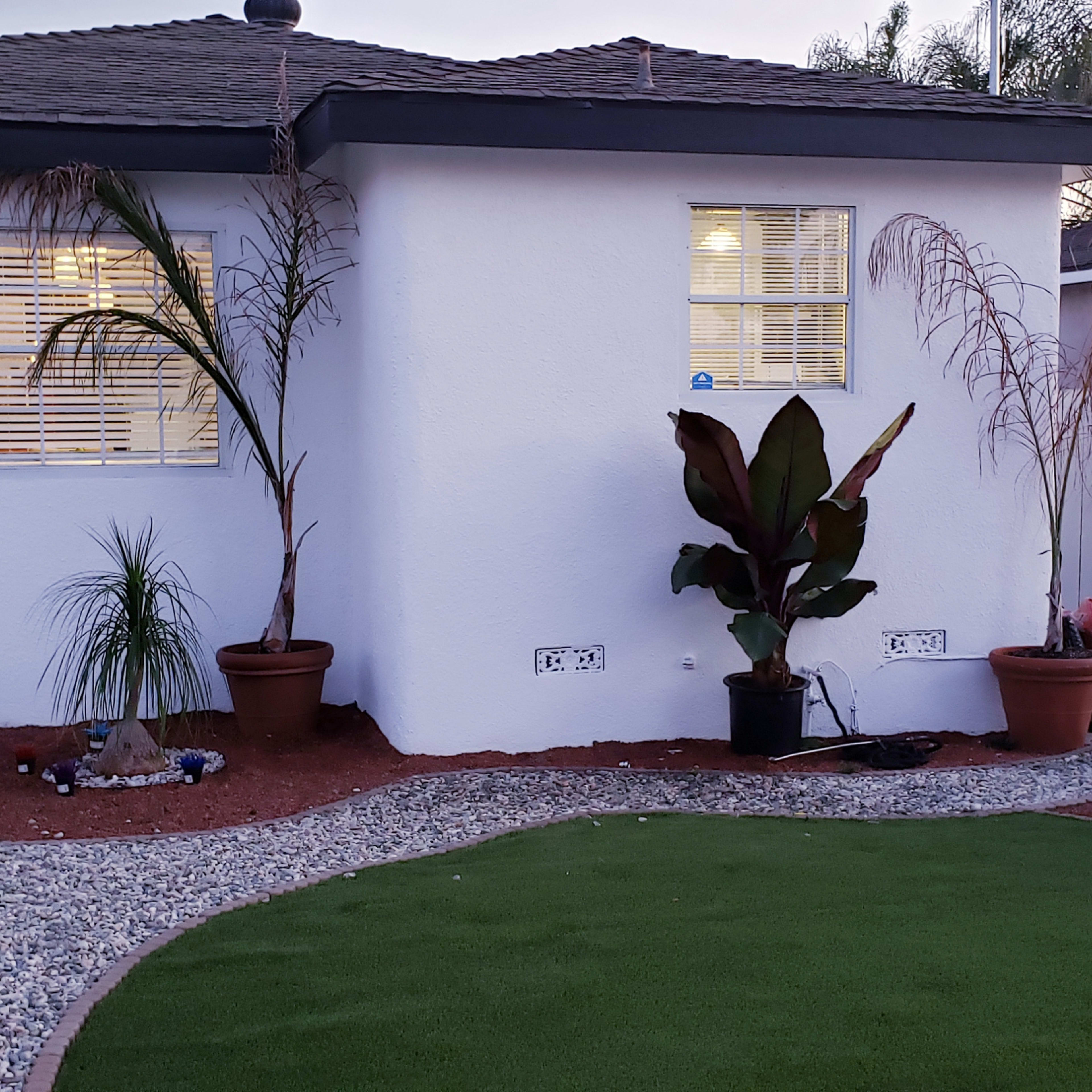 A small front yard with a white house, landscaped area featuring potted plants, gravel, and artificial grass.