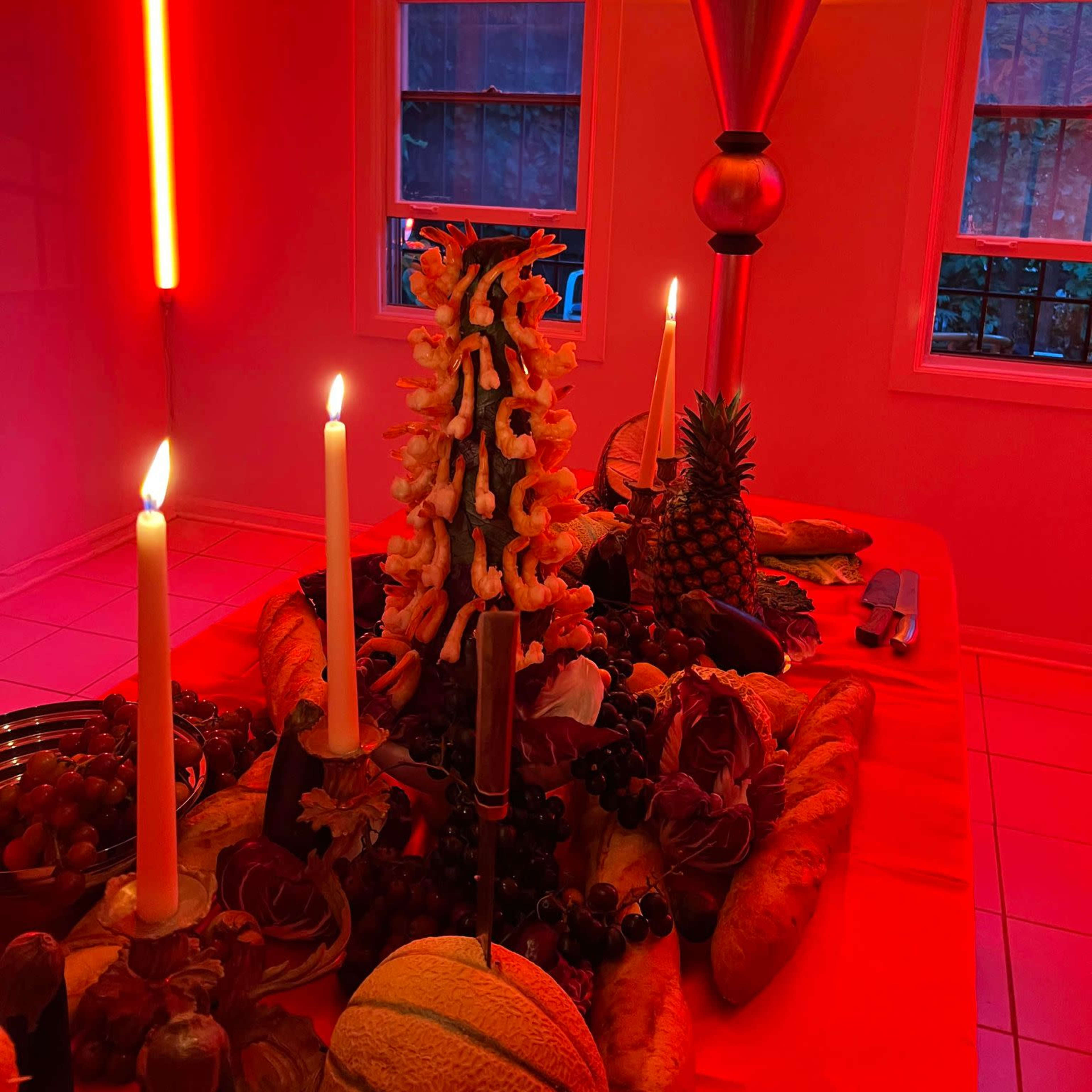 A table is adorned with an assortment of fruits, candles, and a decorative centerpiece, all illuminated by red and orange lighting.