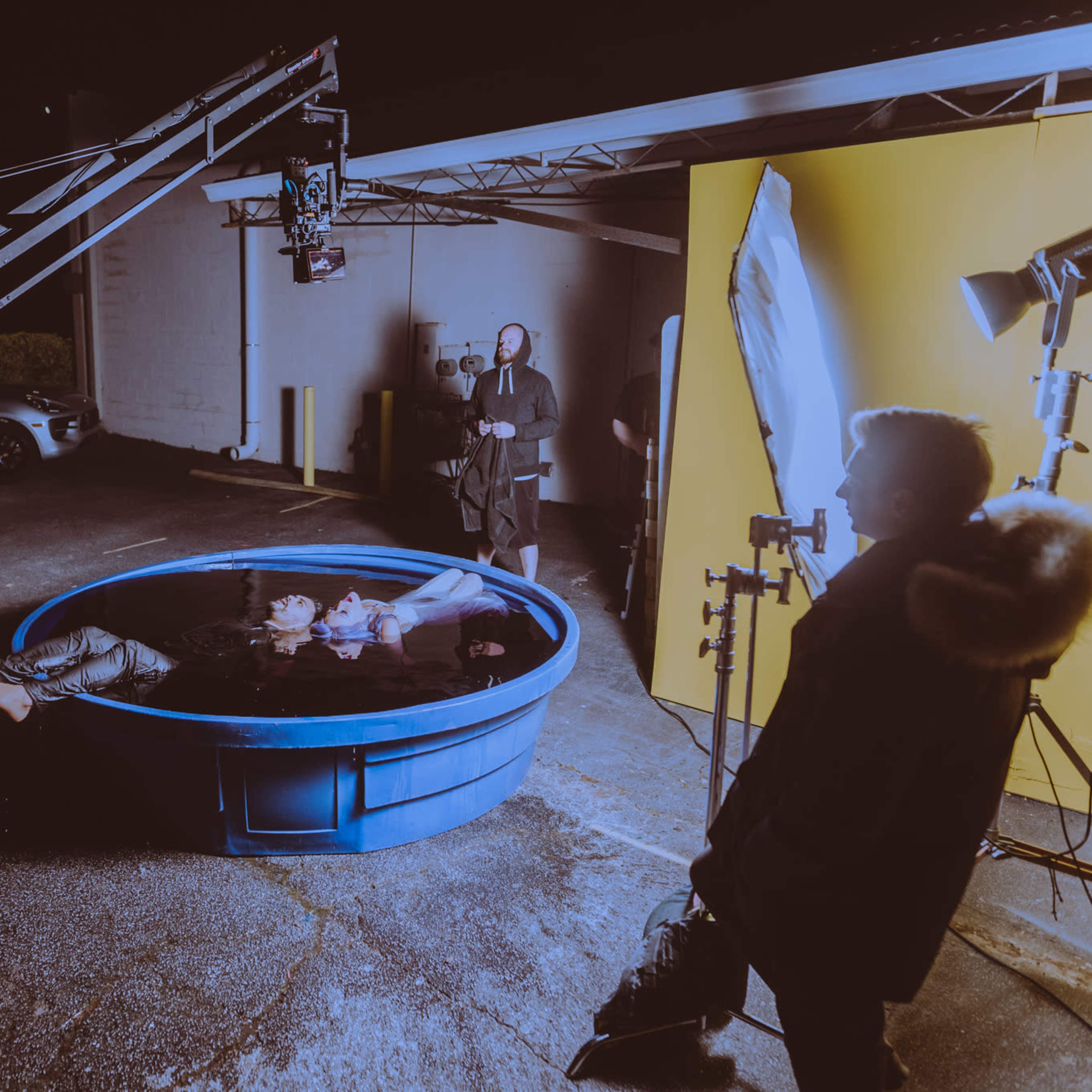 A blue pool is set up outdoors with a camera crane positioned above it, while a crew prepares for a photoshoot at night.
