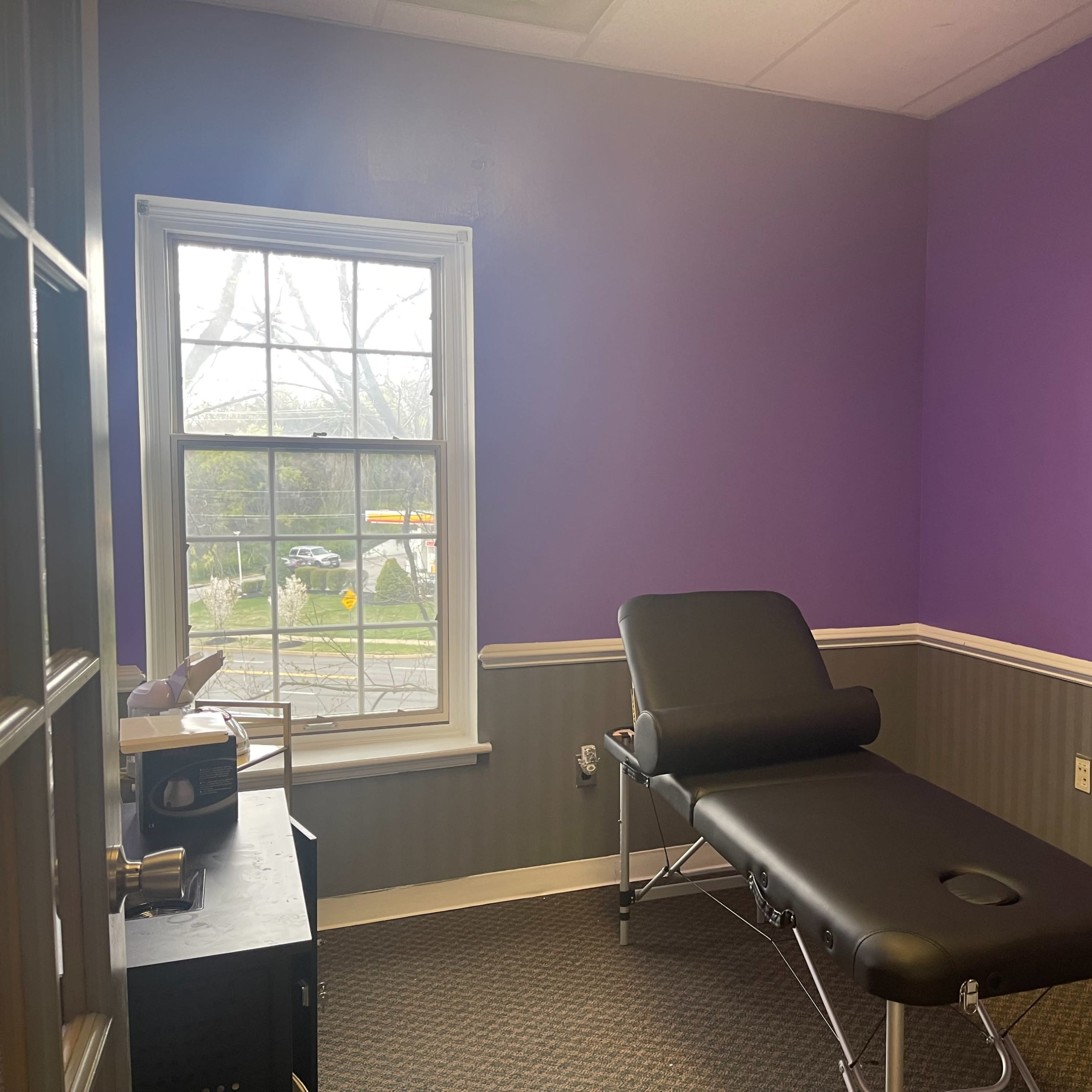 The room features a black treatment table, a small desk, and purple walls with a window showing an outdoor view.